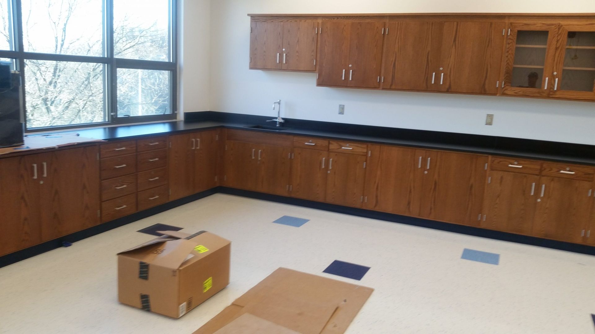 A school lab room with brown cabinets, black countertops, and a window. A cardboard box sits on the floor.