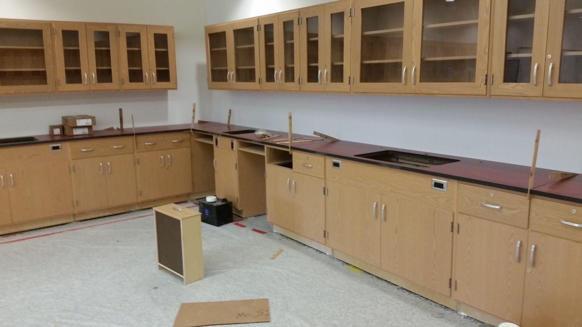 Empty lab with wood cabinets, tan and maroon countertops, and upper glass-front cabinets; under construction.