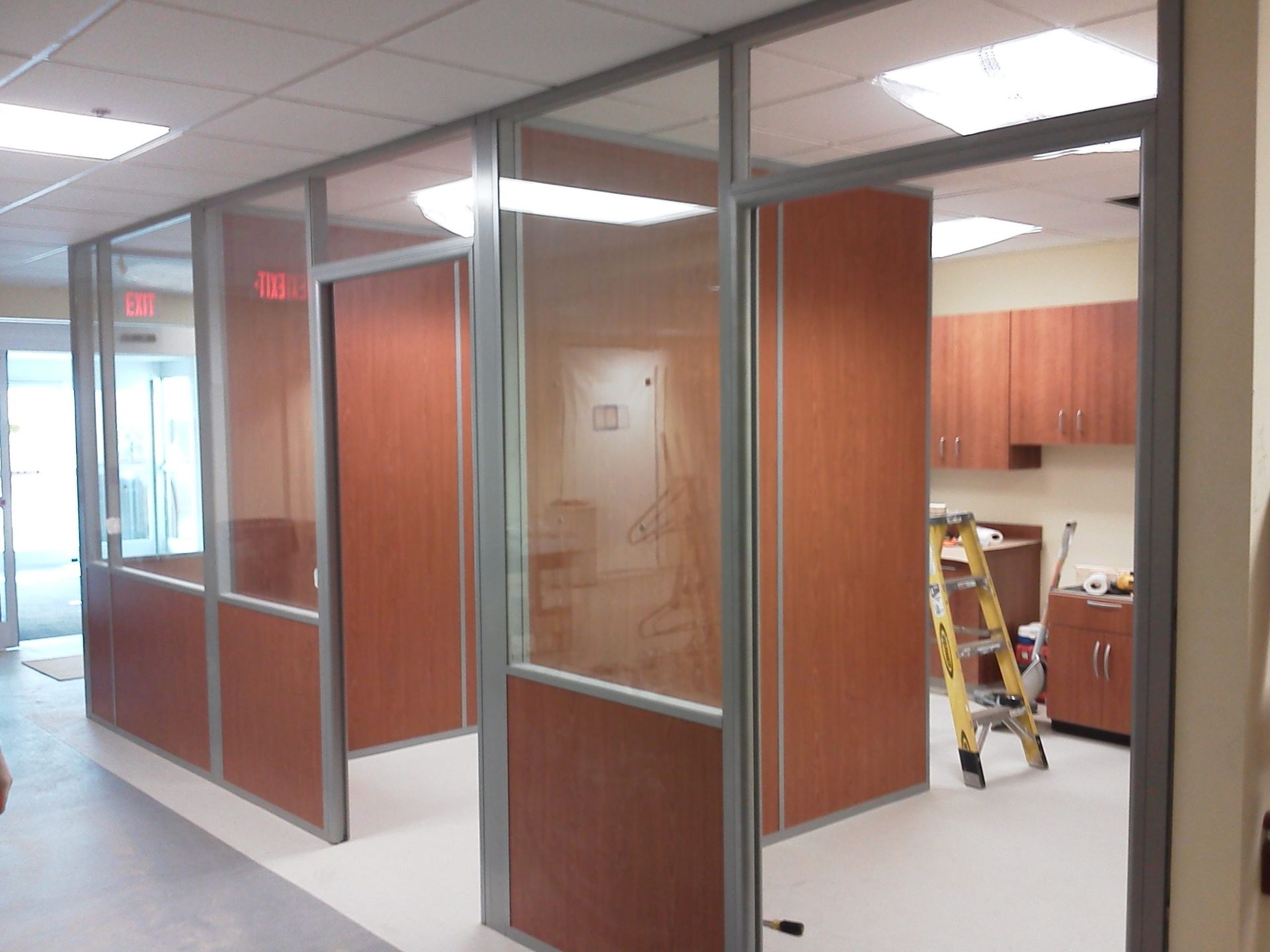 Office cubicles with glass panels and wood-look walls, gray metal frames, under bright fluorescent lights.
