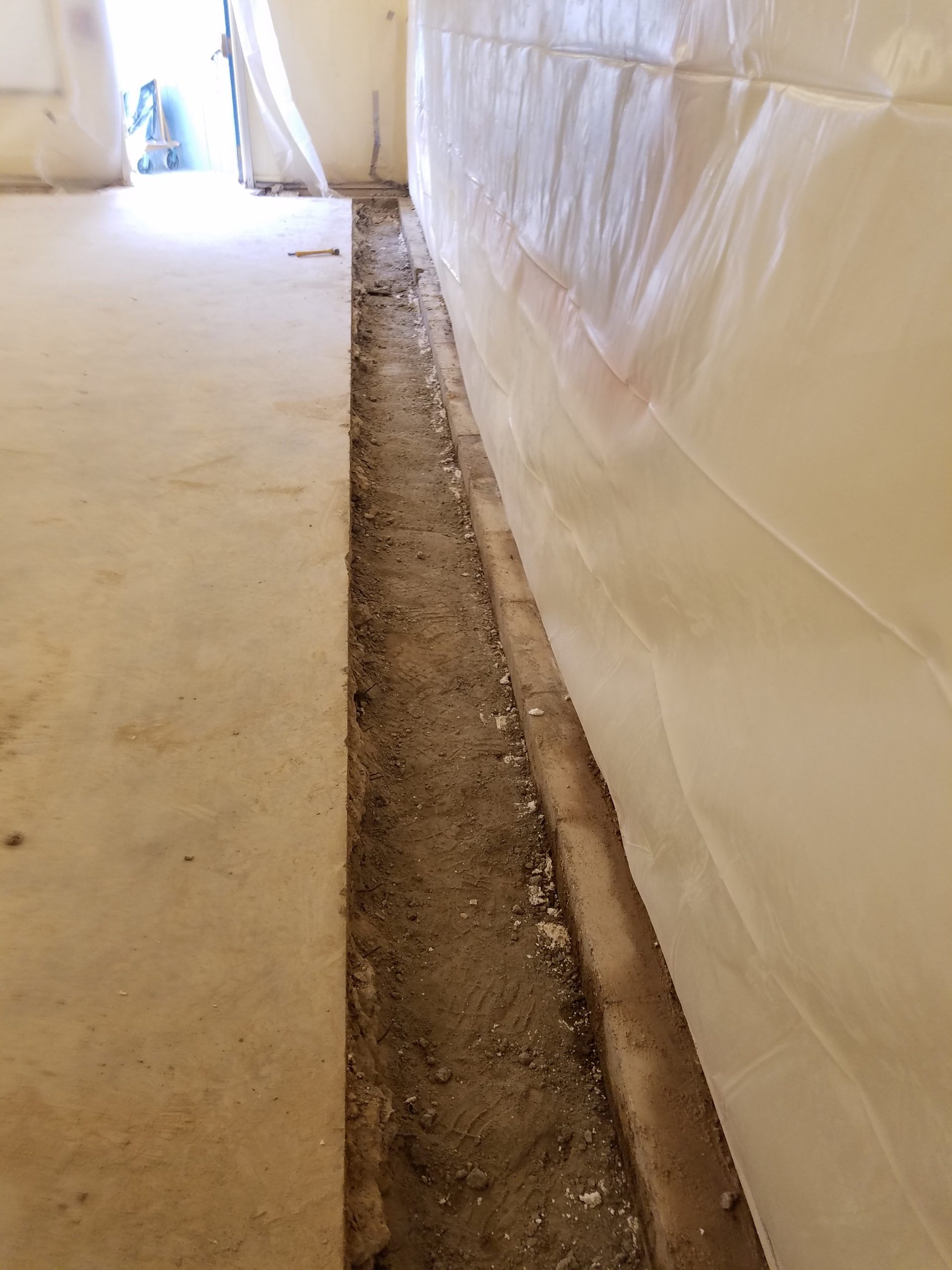 A narrow trench along a wall in a concrete room. Plastic sheeting covers the wall.