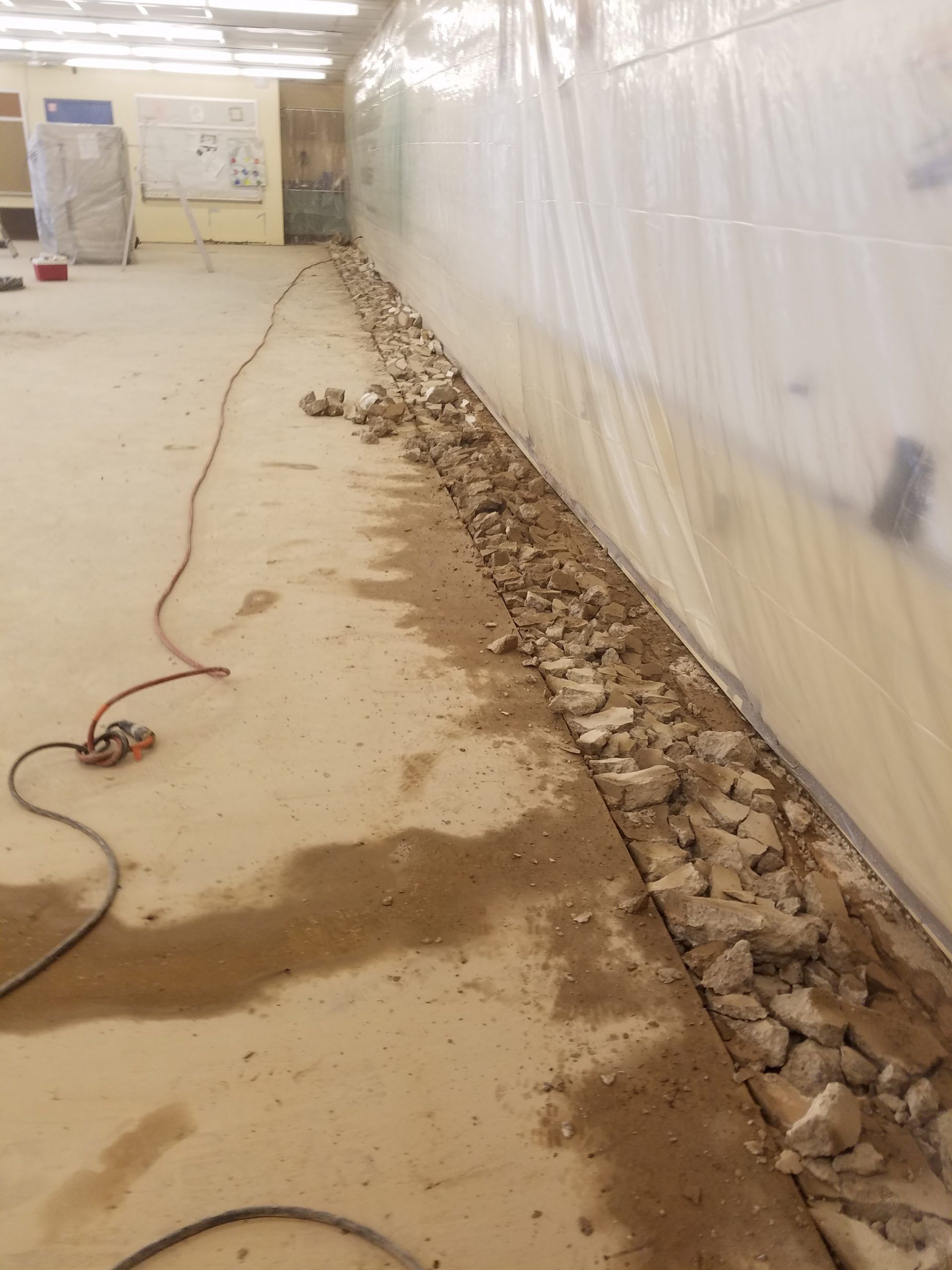 Concrete floor with debris along a wall covered in plastic sheeting; electrical cord on the floor.