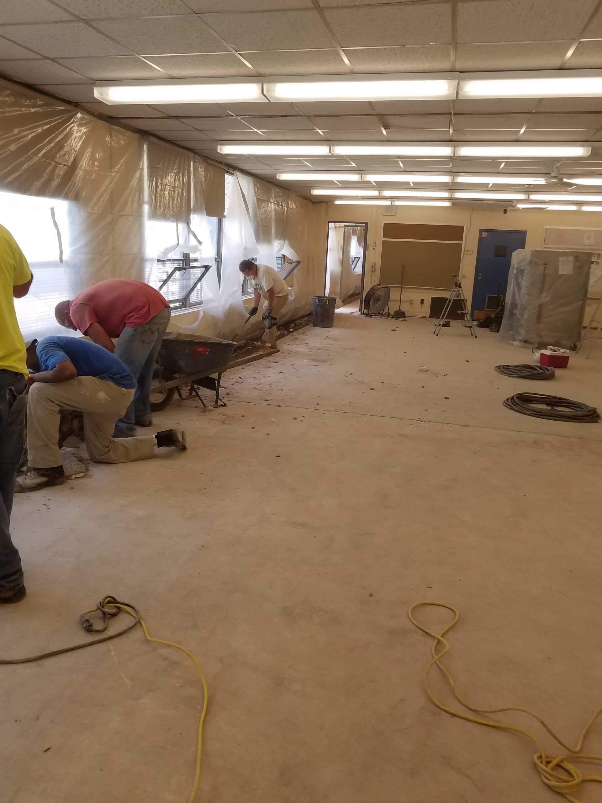 Workers in a dusty room, demolishing a wall. Brown dust, white ceiling lights, tarps, and tools visible.