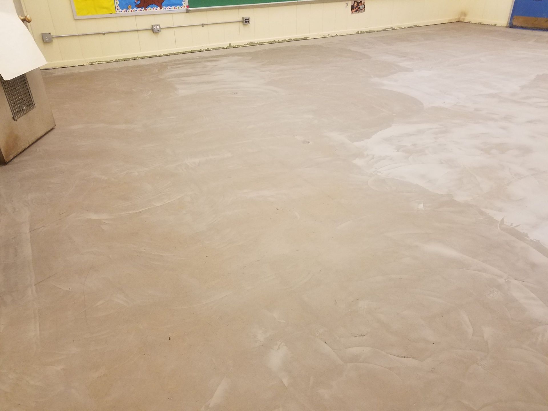 Tan, textured concrete floor in a room, with a partially visible wall and a small radiator on the left.