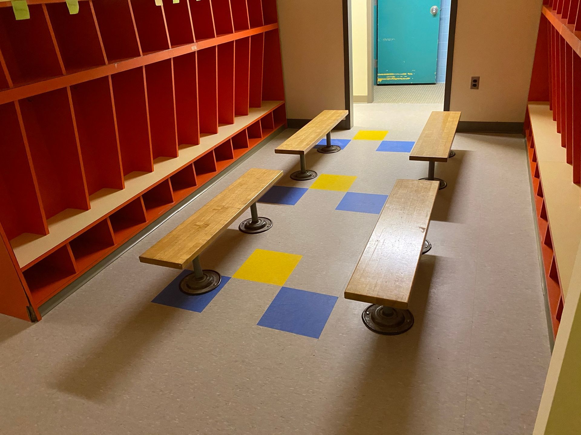 Red lockers and benches in a hallway with blue and yellow floor tiles, leading to a doorway.