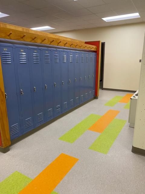Blue lockers line a hallway with colorful floor accents. Wooden beam overhead, red door at the end.