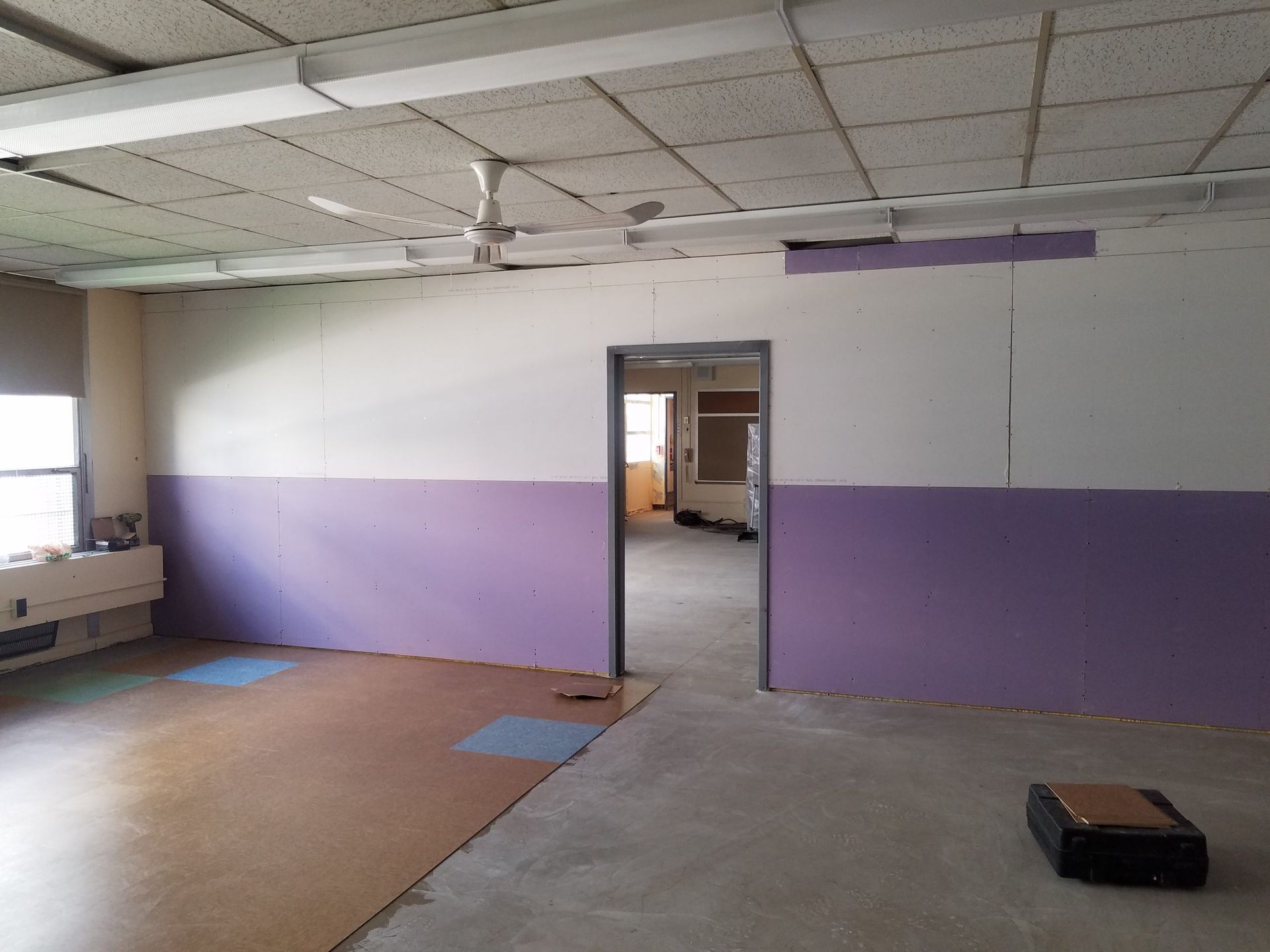Empty room with purple and white walls, door, and ceiling lights; floor is part carpet, part concrete.