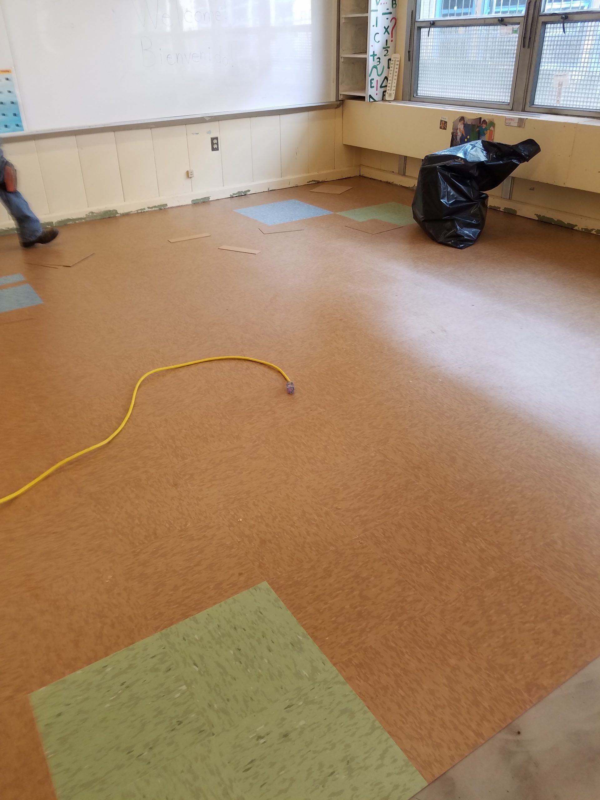 Brown and green floor with exposed glue patches in a classroom; yellow cord, trash bag, and person's leg visible.