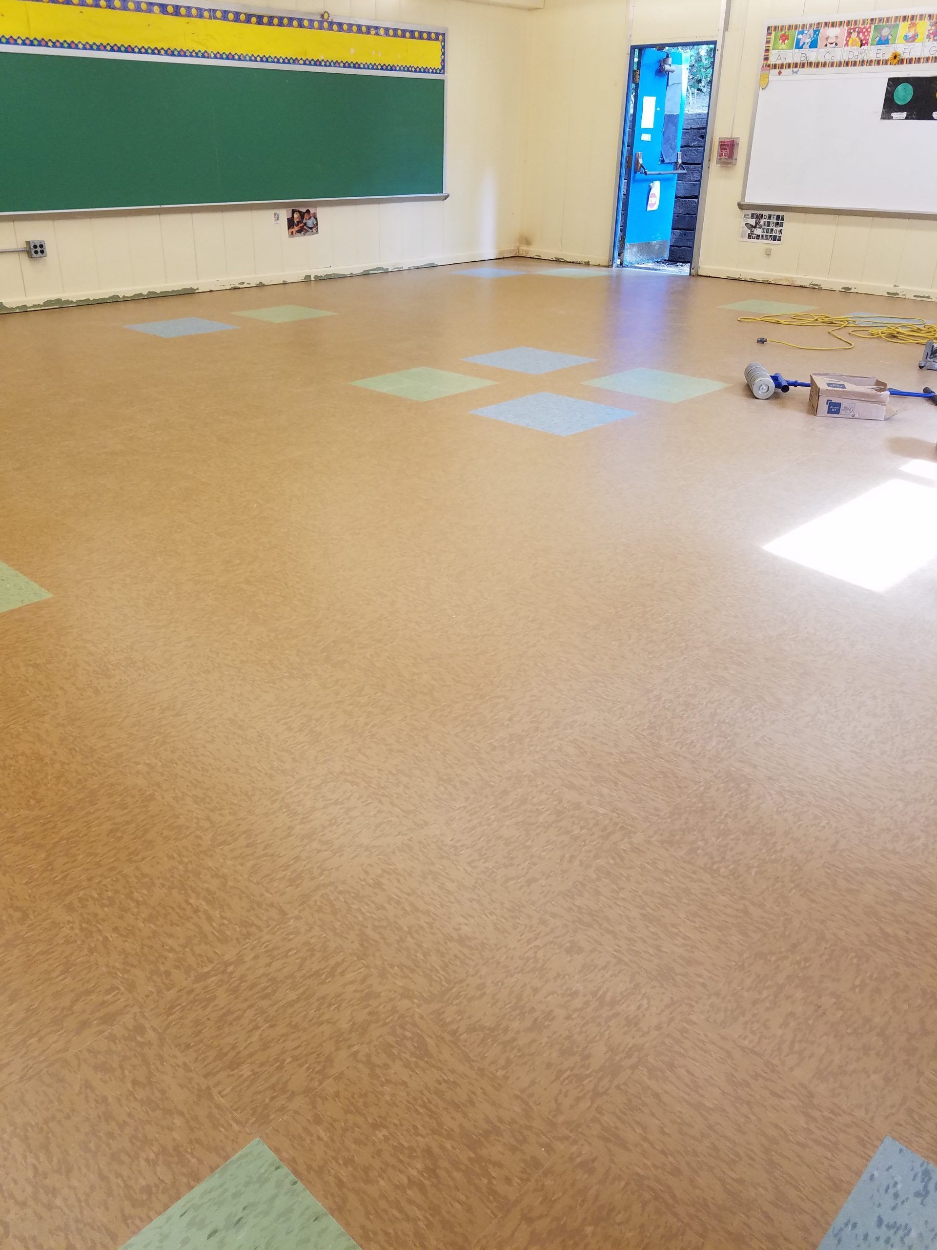 Empty classroom with tan speckled floor and colorful squares. Blackboard and whiteboard visible.