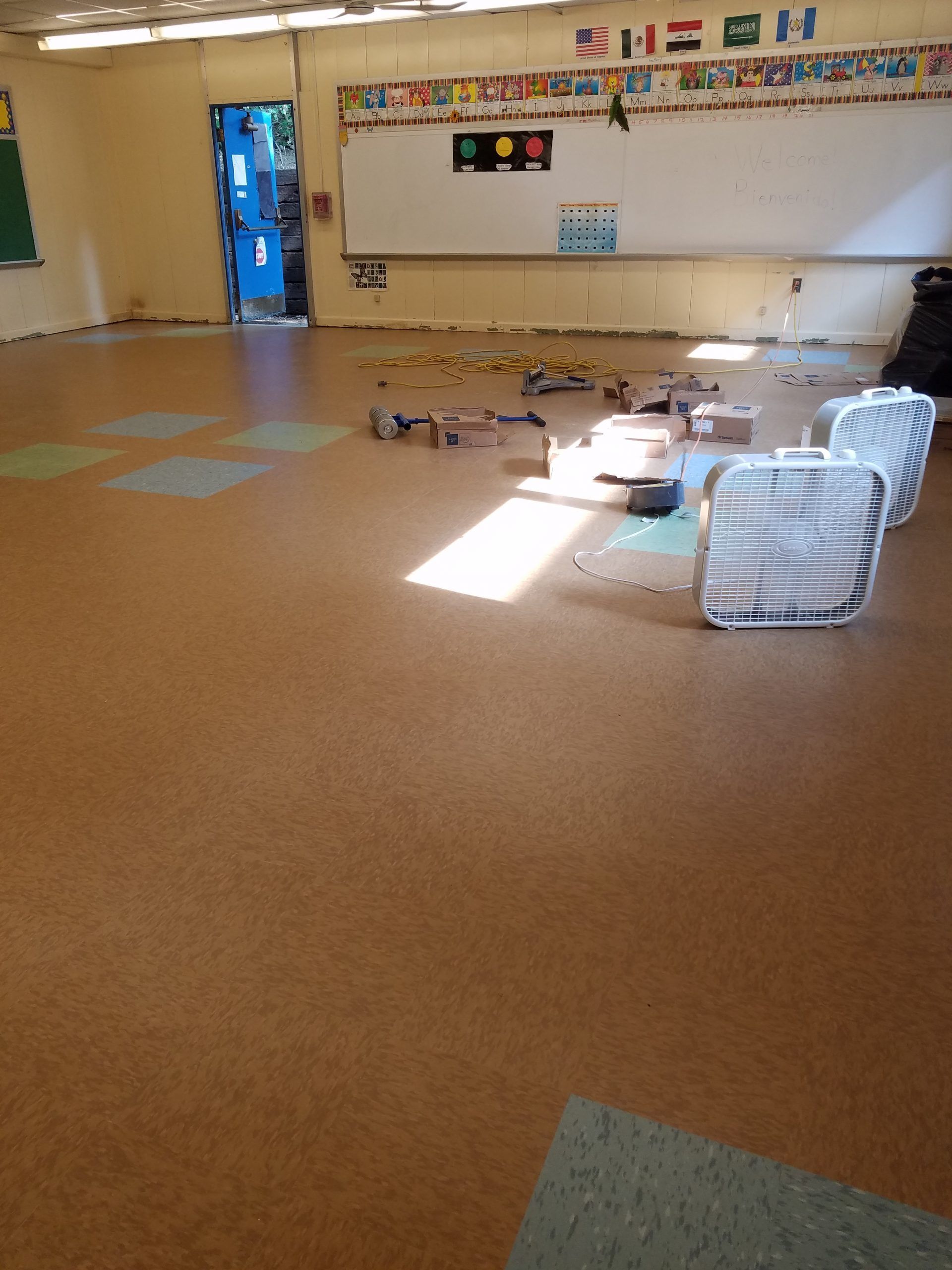 Empty classroom with brown floor, whiteboard, and scattered toys. Two fans sit near the center.