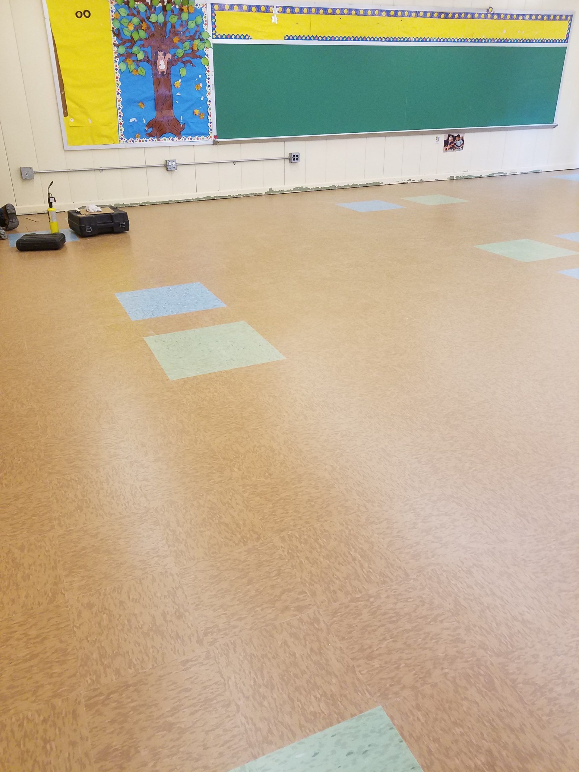 Classroom with brown floor and green and blue squares. Green chalkboard, bulletin board.