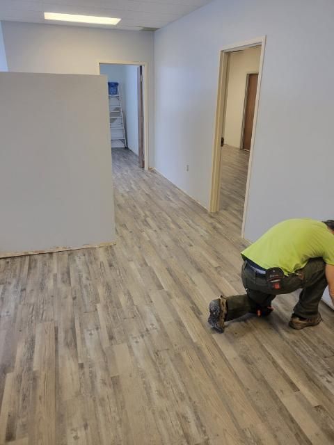 Man installing light gray wood-look flooring in a room with white walls and open doorways.
