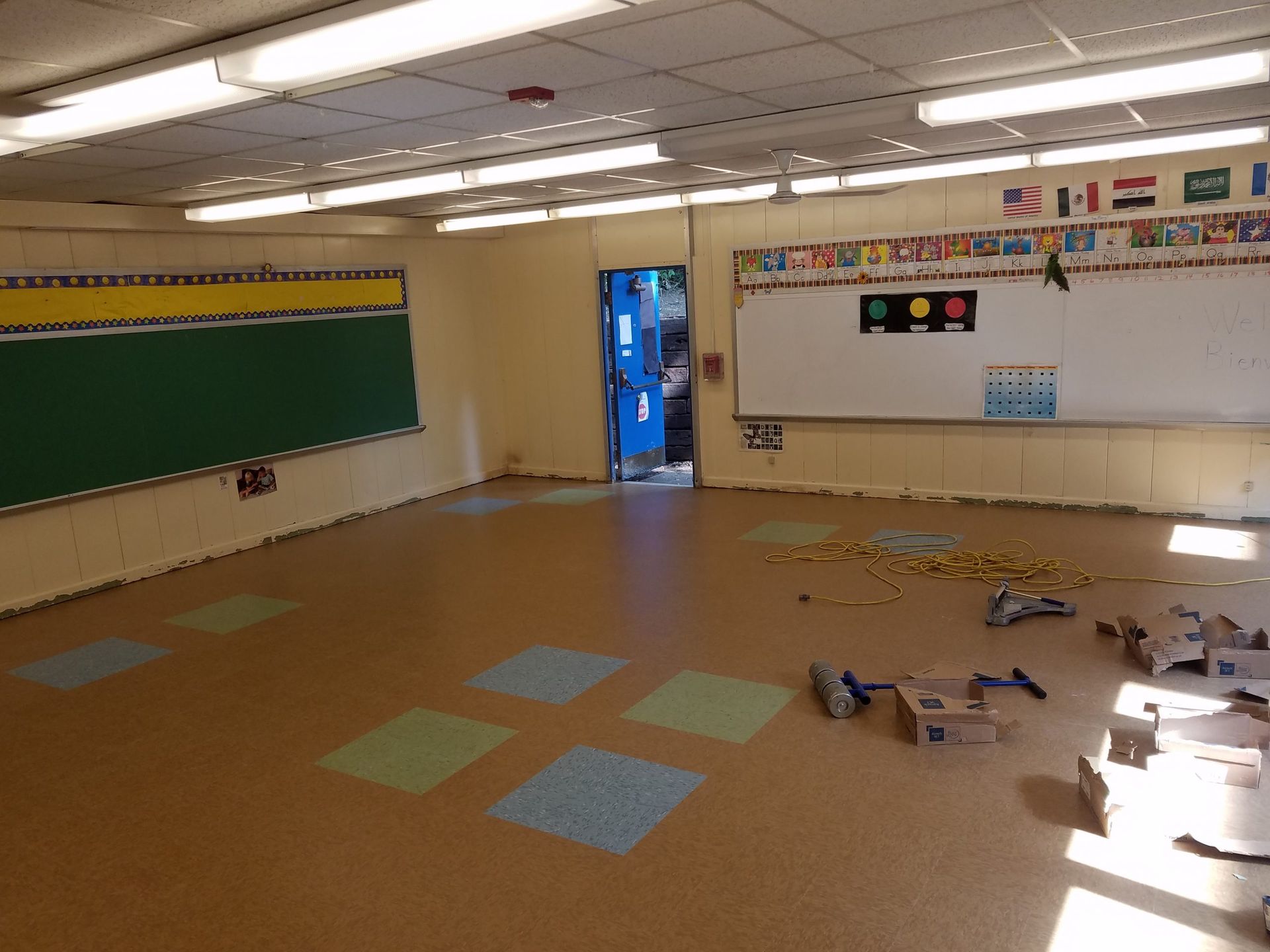 Empty classroom with green chalkboard, white board, colorful floor tiles, and open door.