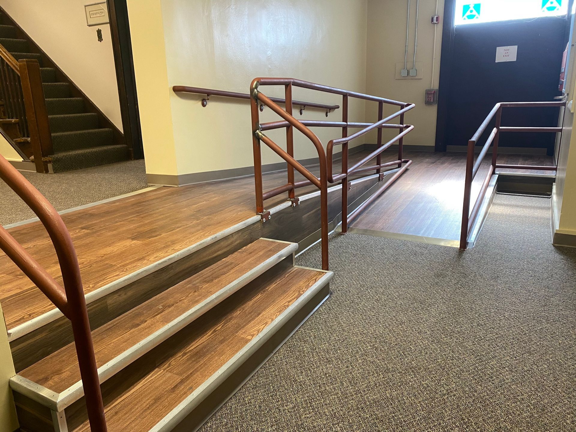 Ramp and stairs in a building entrance. Brown wooden ramp and steps, brown handrails. Door at top, stairs to the left.