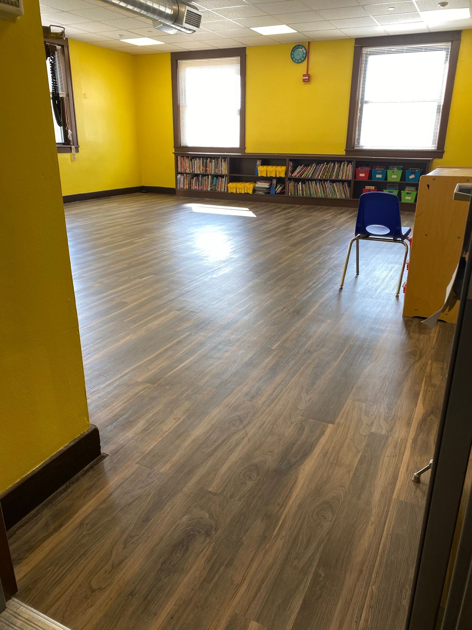 Empty yellow classroom with wood-look floor, windows, bookshelves, and a blue chair.