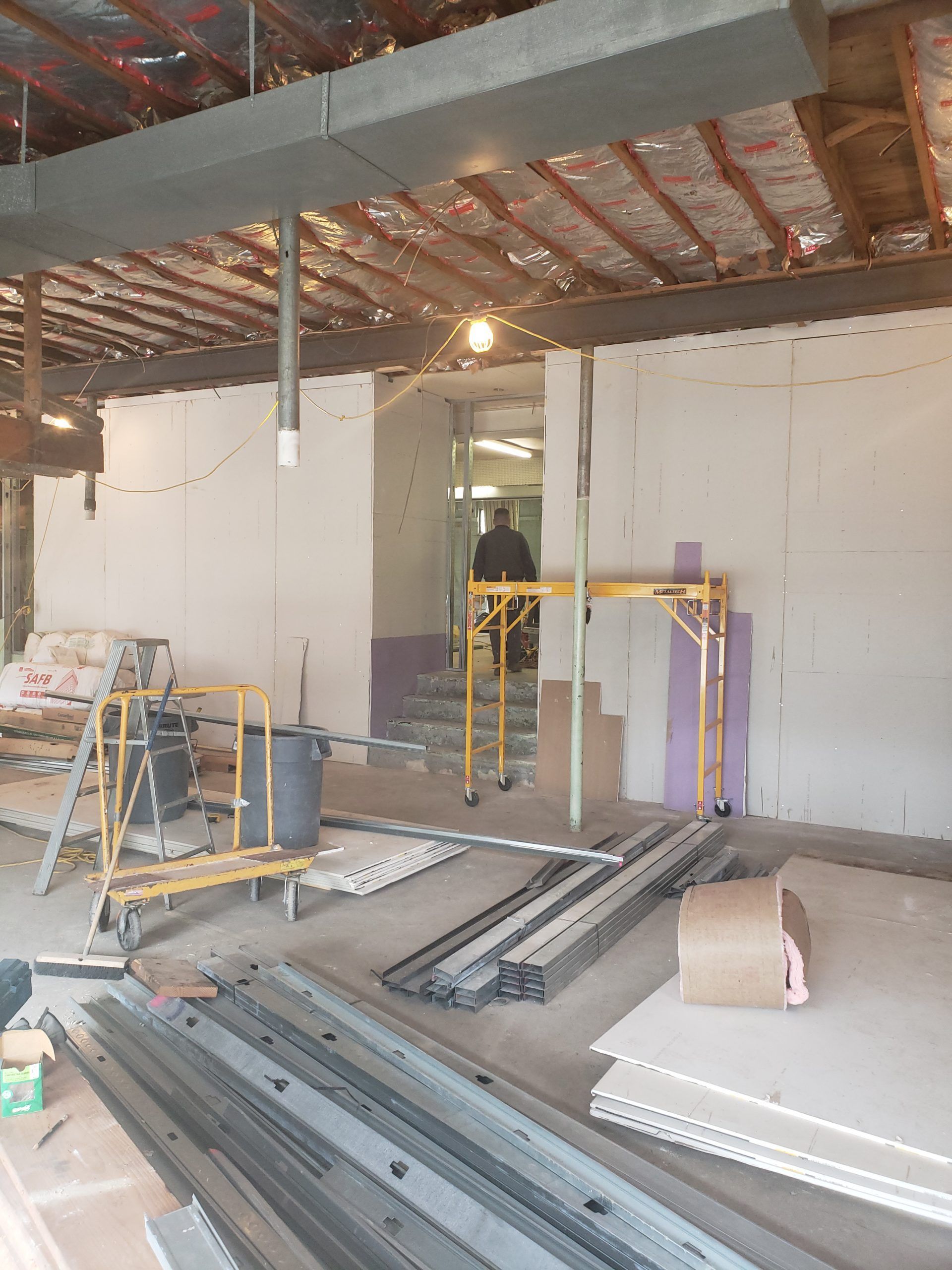 Construction site interior with drywall, scaffolding, and materials. Person in doorway.