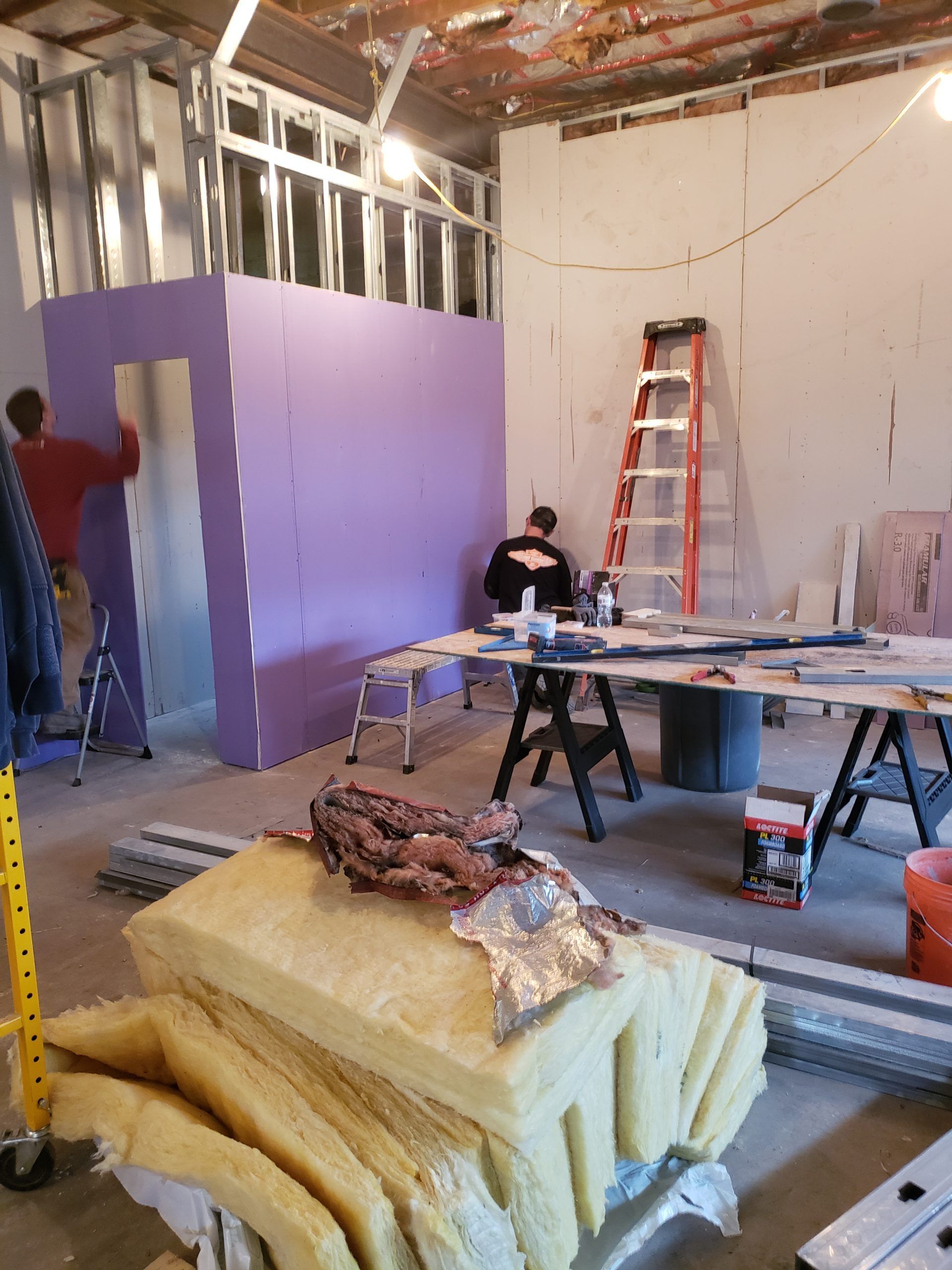 Construction site interior. Workers installing purple drywall, one on a ladder. Insulation and tools are visible.