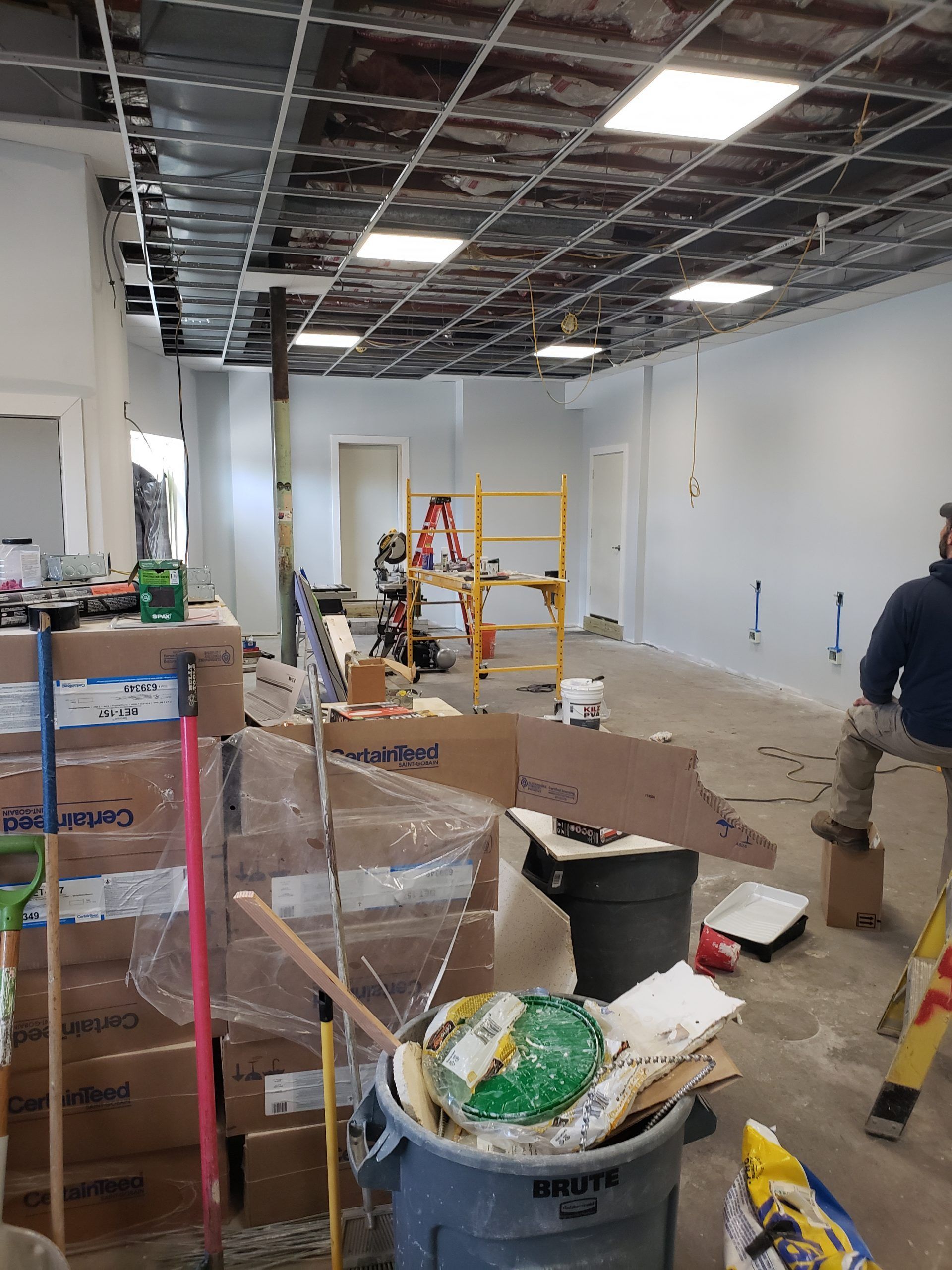 Interior construction site with boxes, tools, and a worker. Exposed ceiling, white walls, and debris on the floor.