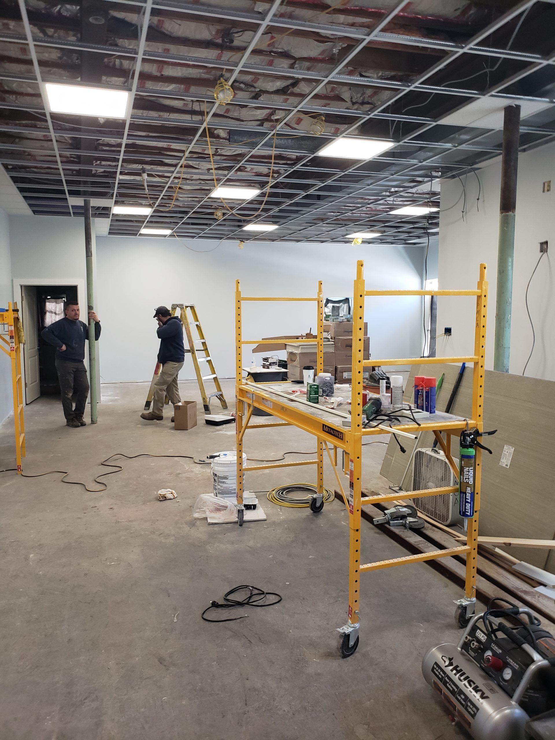 Construction site interior: workers, scaffolding, exposed ceiling grid, concrete floor, tools.