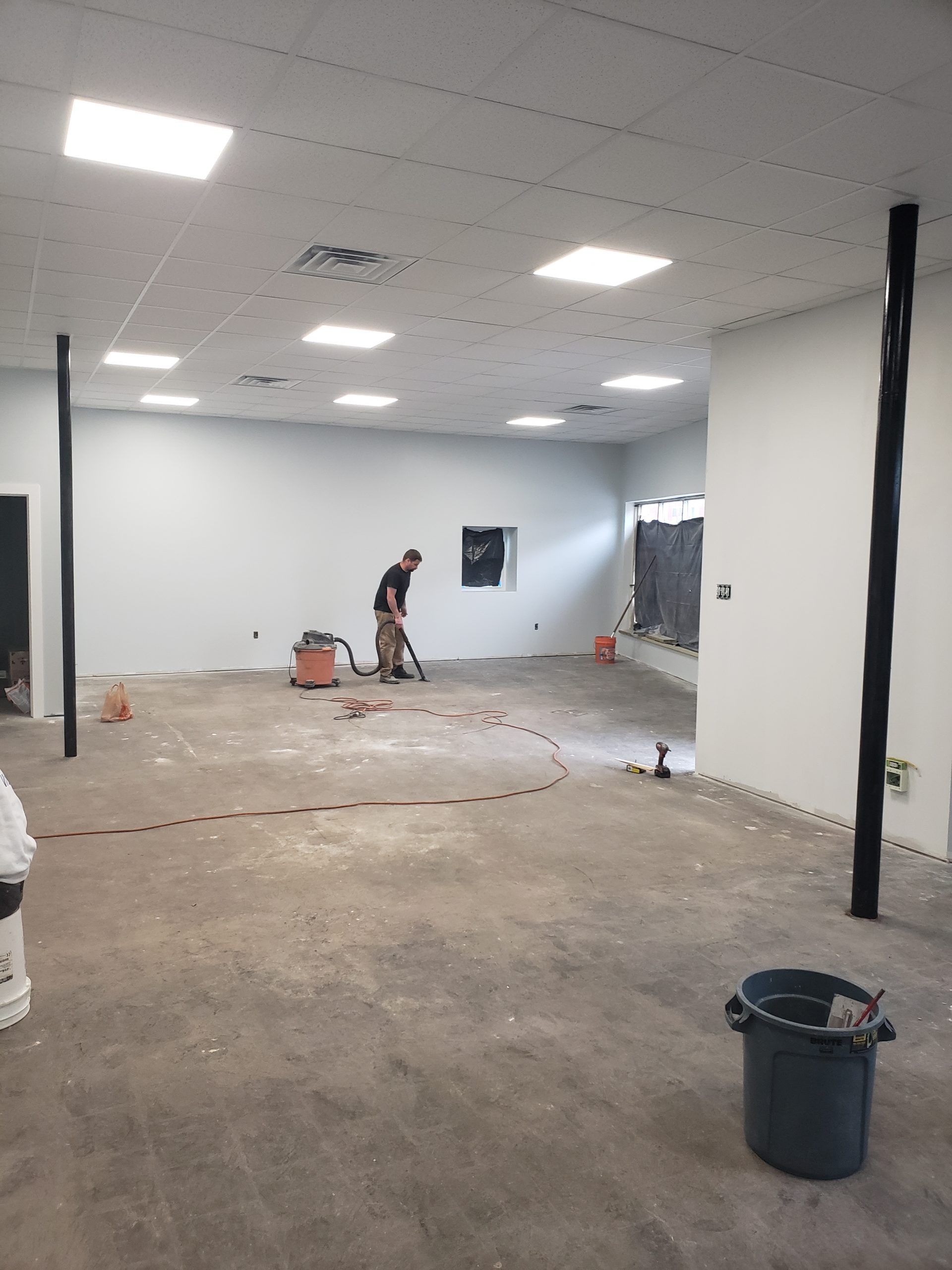 Interior of a construction site. A worker vacuums the concrete floor. White walls, black support columns, and overhead lights.
