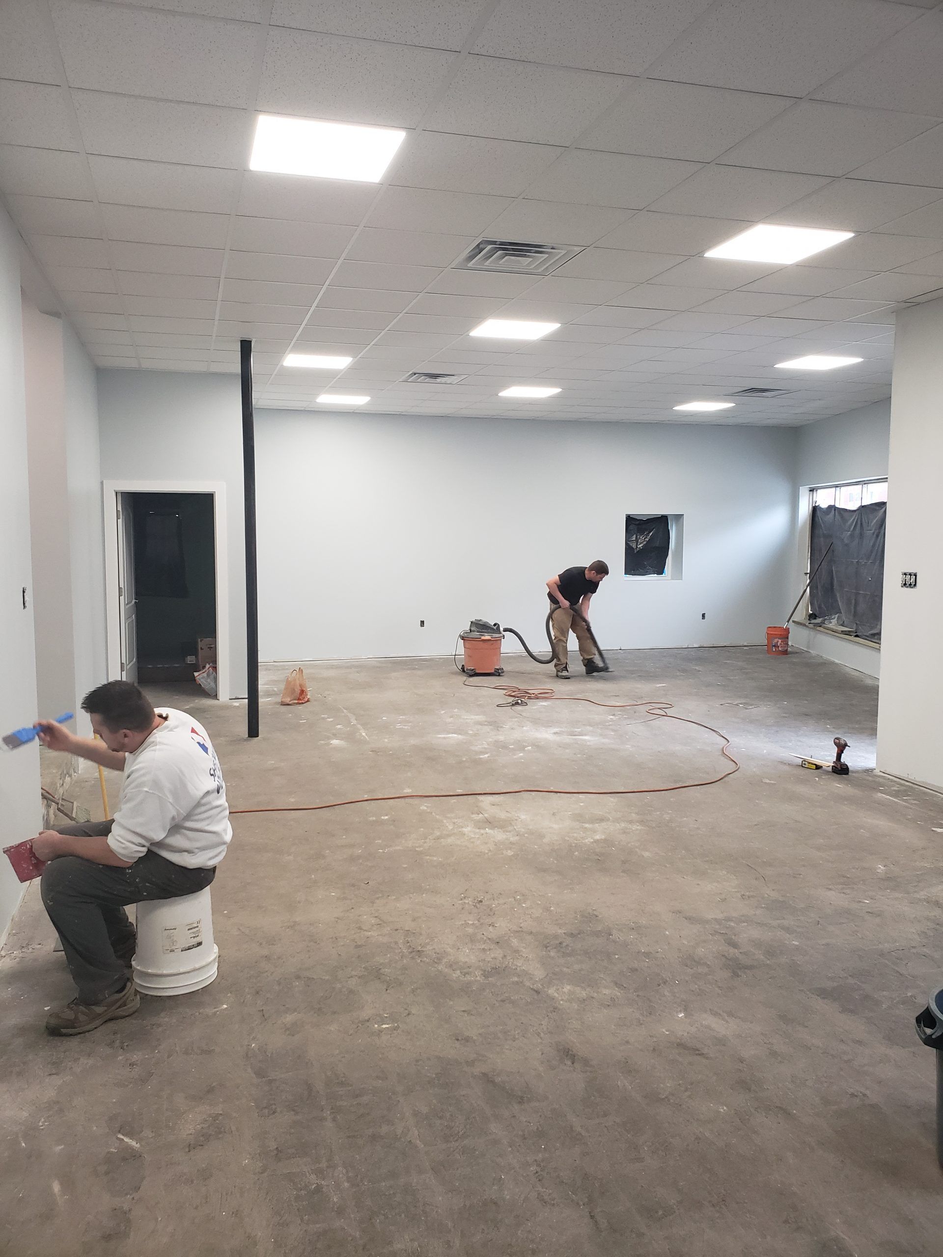 Workers inside a room with concrete floor, drywall walls, and overhead lights; one is seated, and another is using a power tool.