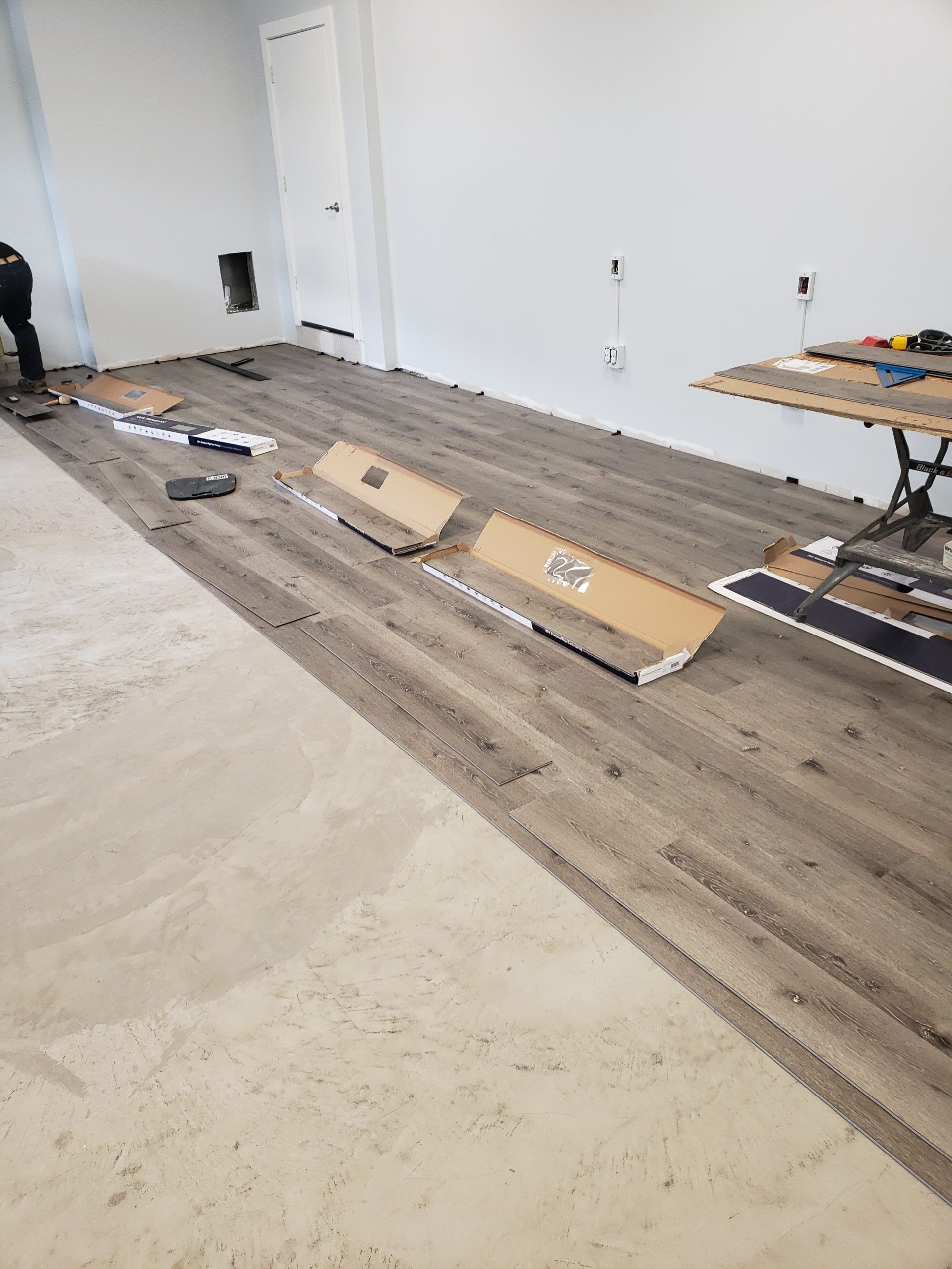 Installing wood-look flooring on a light concrete floor. Flooring material, tools, and partial wall visible.