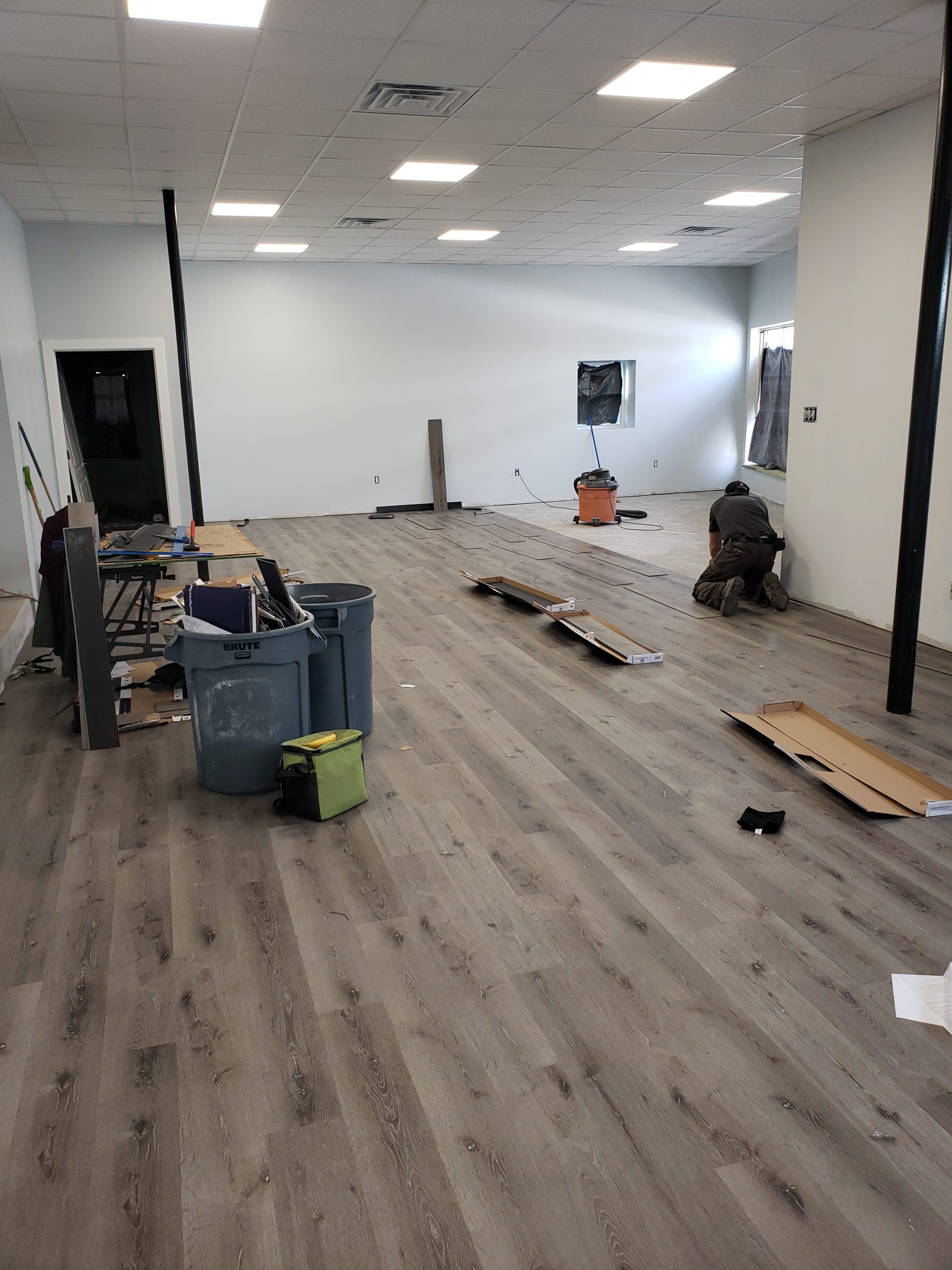 Interior of a room under construction with gray flooring. A worker kneels. Tools and debris scattered.