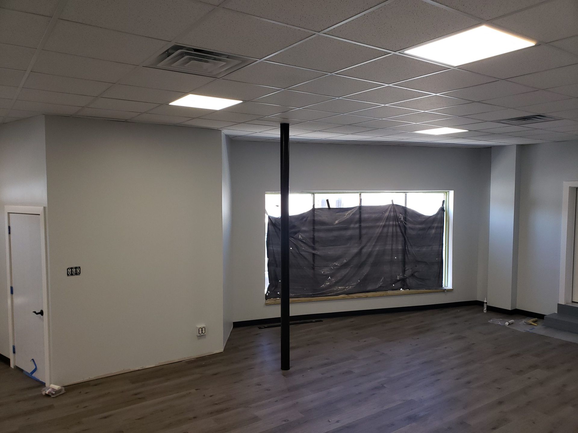 Empty room with gray walls, wood-look flooring, and a central black support beam. Window covered with black tarp.
