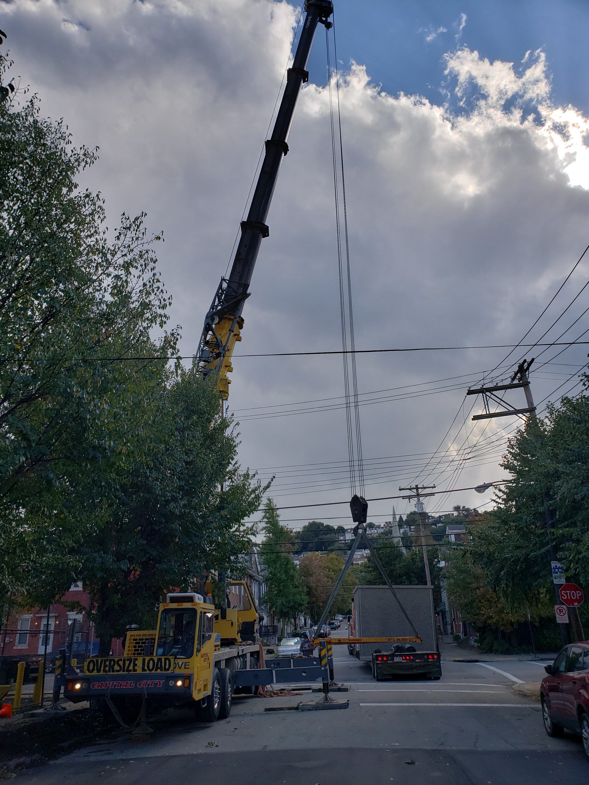 A large crane lifting something in a city street. Utility lines and vehicles are visible, under cloudy sky.