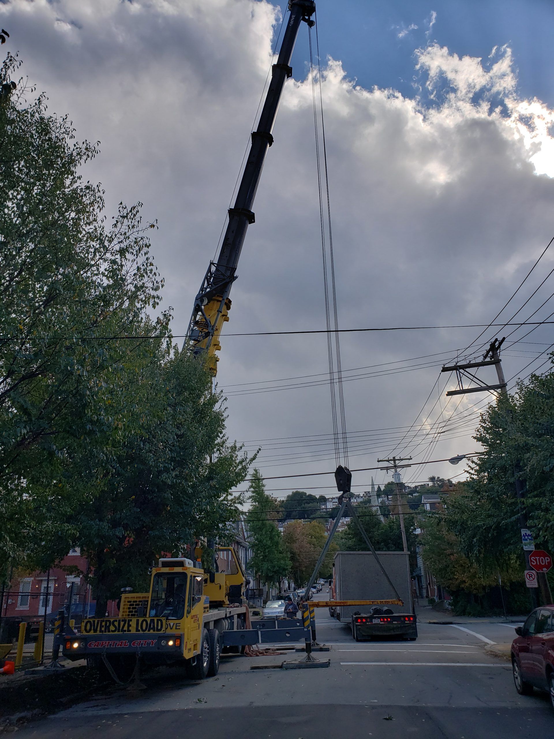 Crane lifting object over a road with utility lines. Trucks and workers are present, overcast sky.