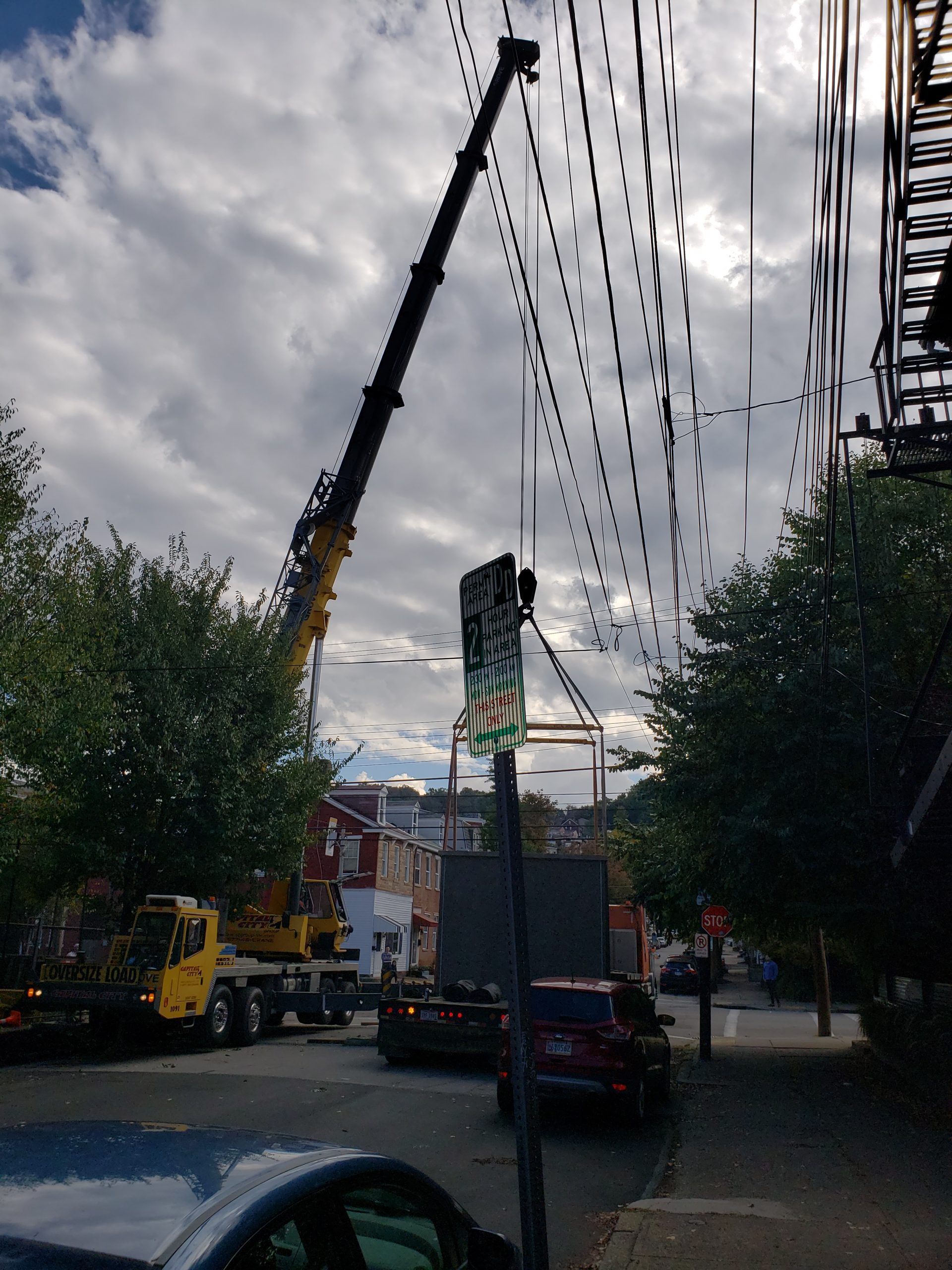 Crane on city street, next to power lines. Vehicles and trees are also present.