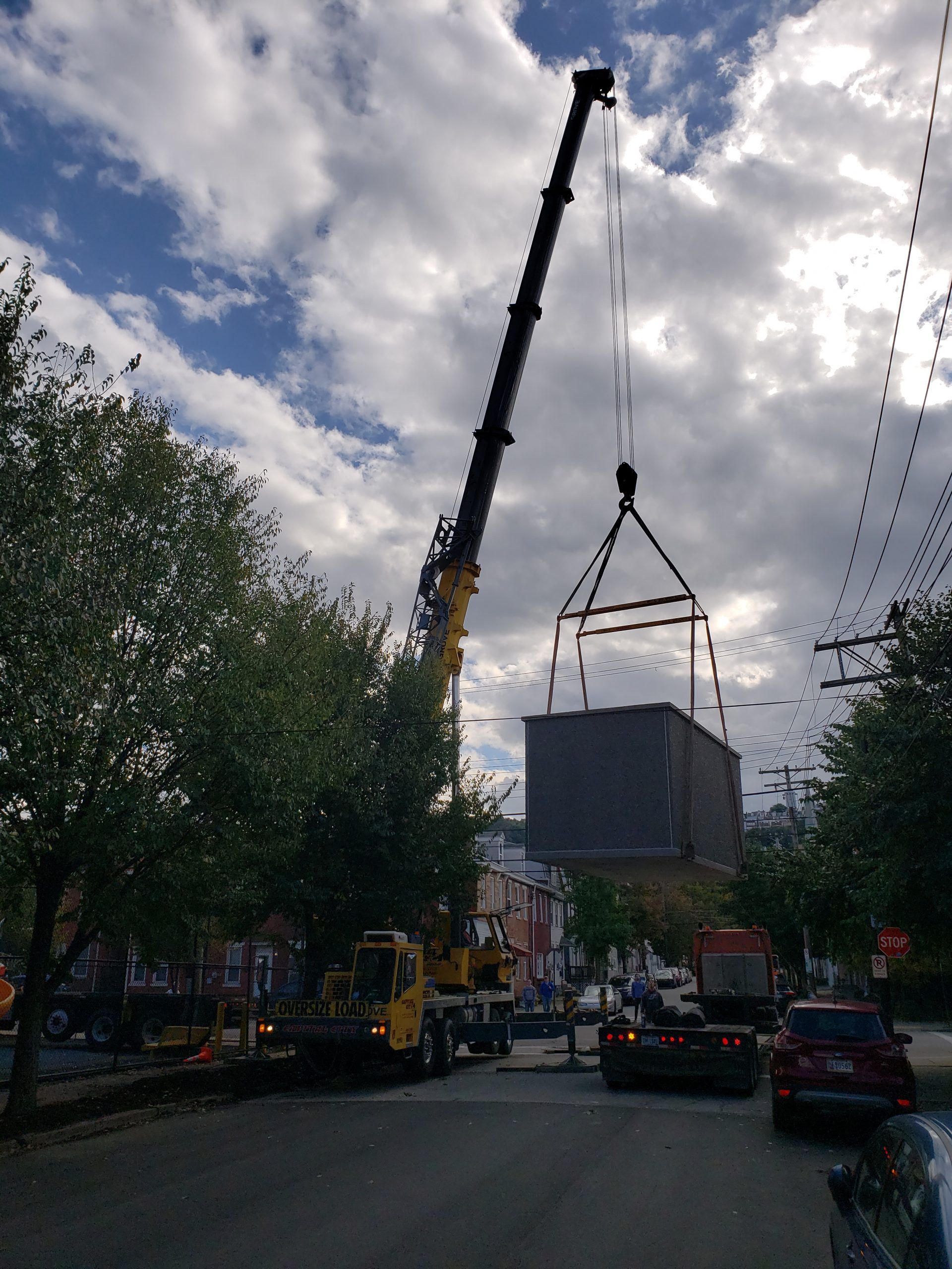A crane lifting a large, square structure over a street, cloudy sky overhead.