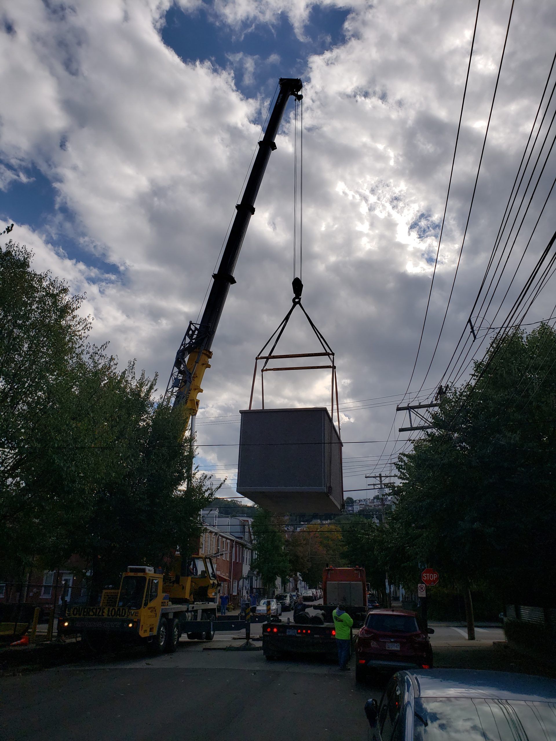 Crane lifting a large gray box structure over a street lined with cars and buildings on a cloudy day.