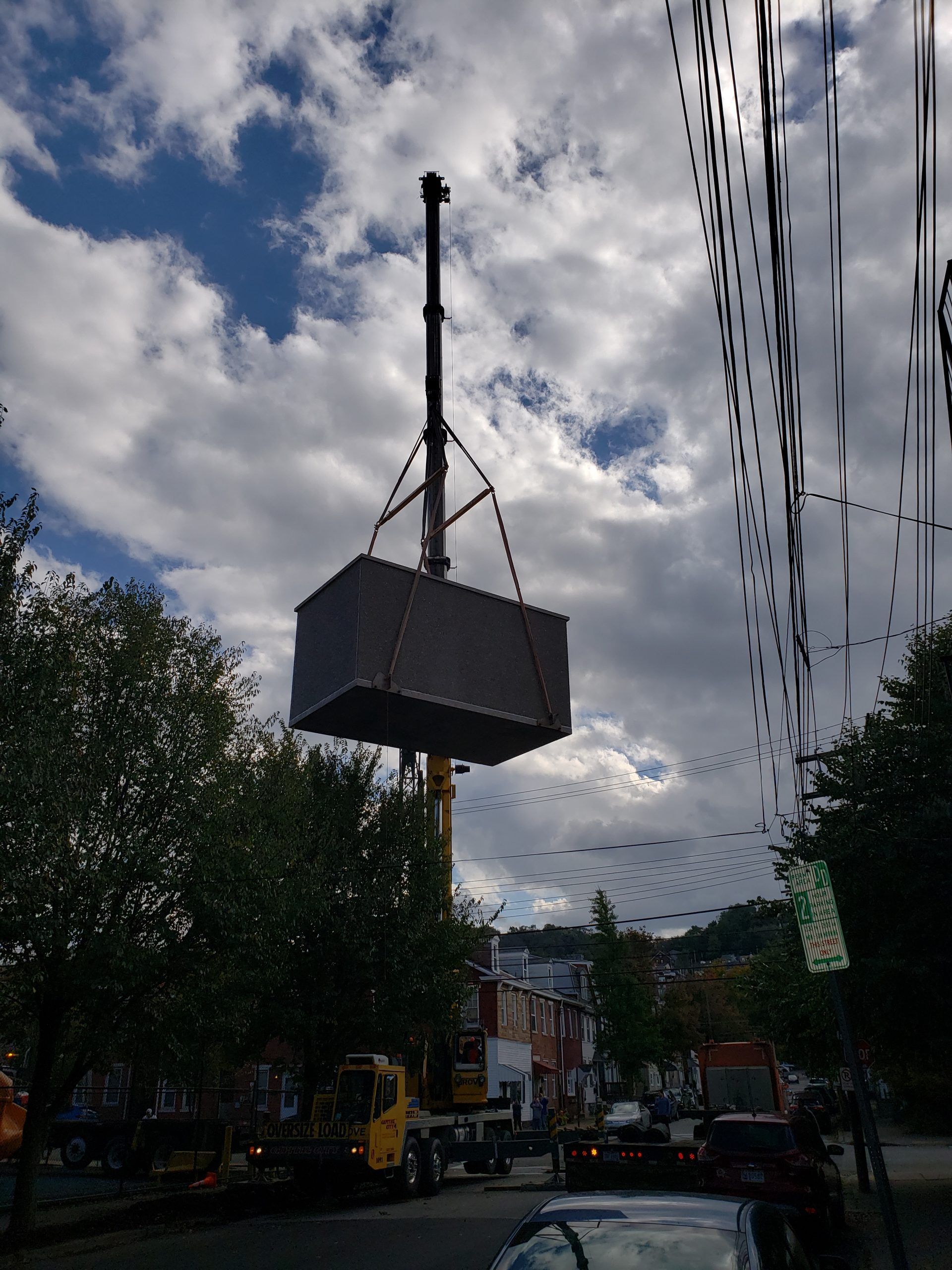 A crane lifts a large, dark container in a city street, under a cloudy sky.