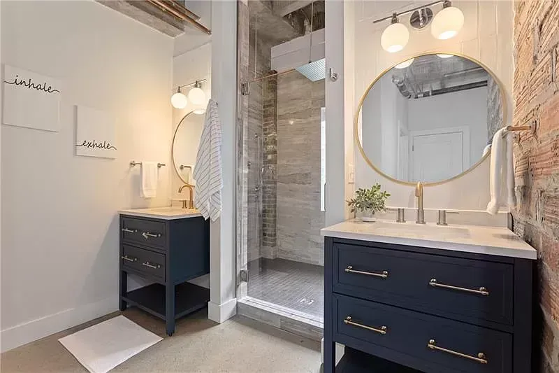 Two navy vanities in a bathroom; one beside a shower, with brick and white walls and gold accents.