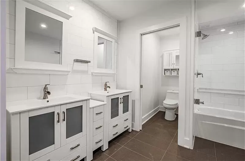 White bathroom with double vanity, toilet, and shower/tub. Brown floor.