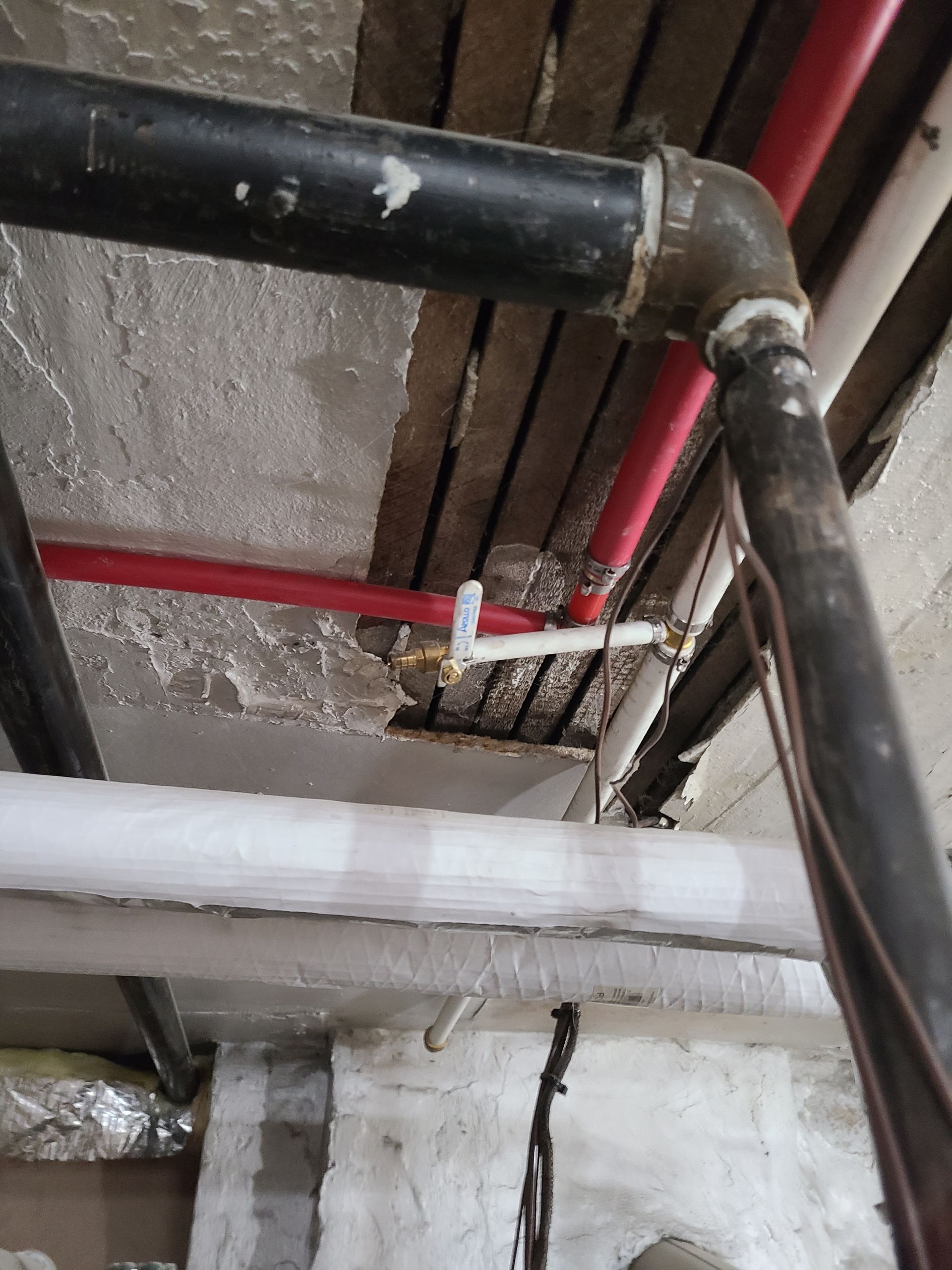 Pipes and plumbing, including red and black pipes, in a basement. Some pipes are connected with brass fittings.