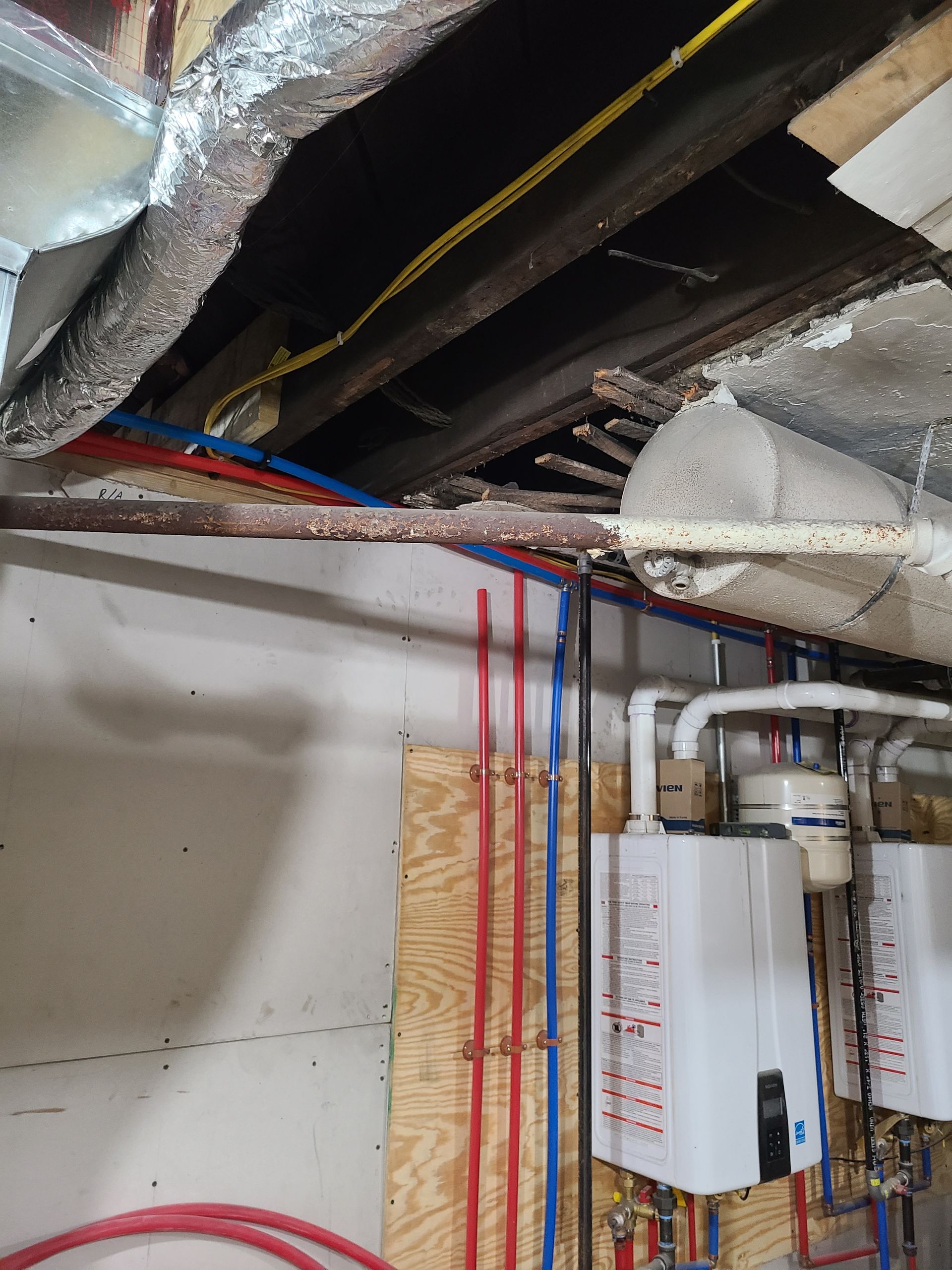 Pipes and wires run along a ceiling with exposed wooden beams, near two white tankless water heaters.