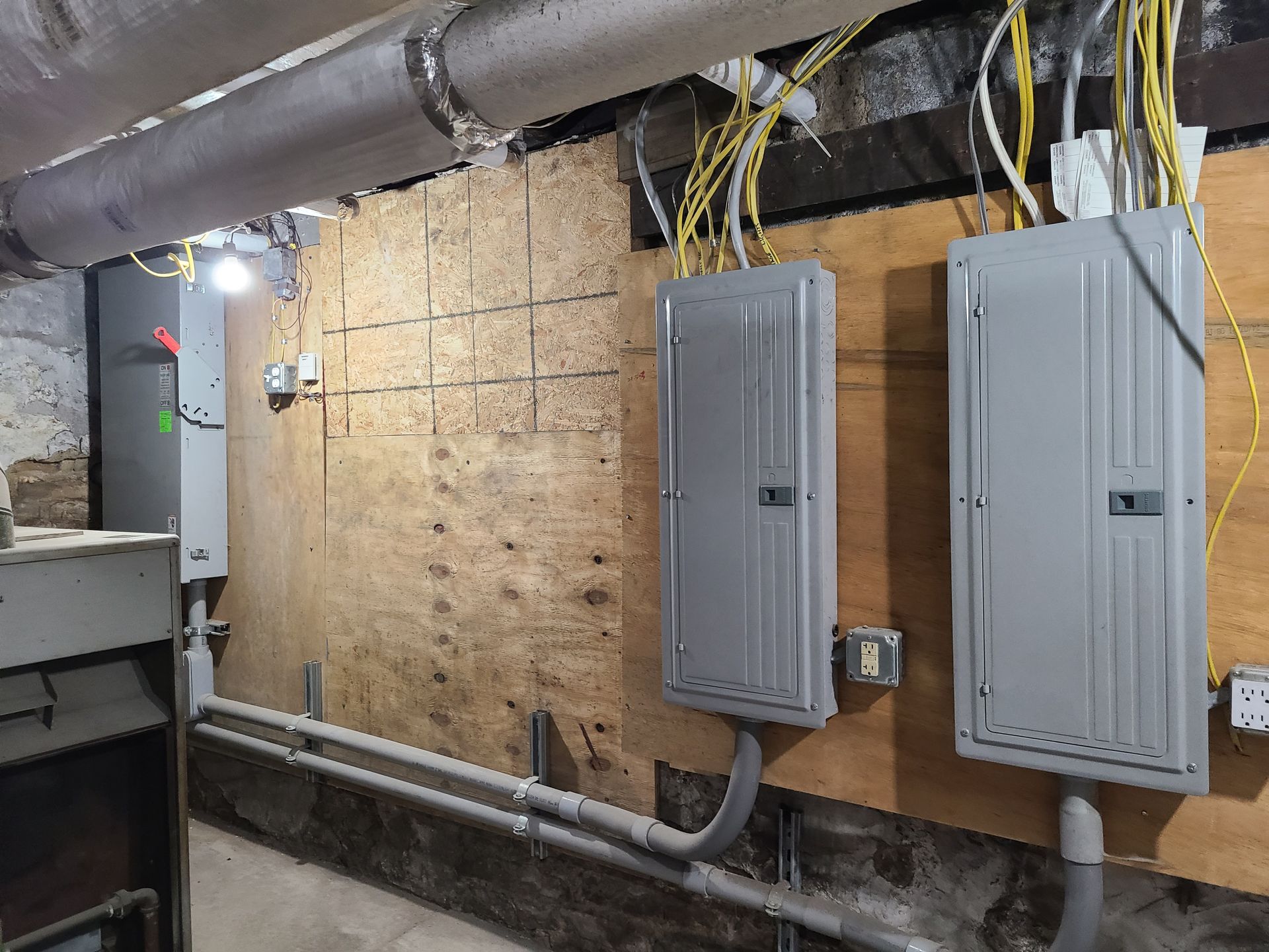 Electrical panels and conduits mounted on a wooden wall in a basement or utility room.