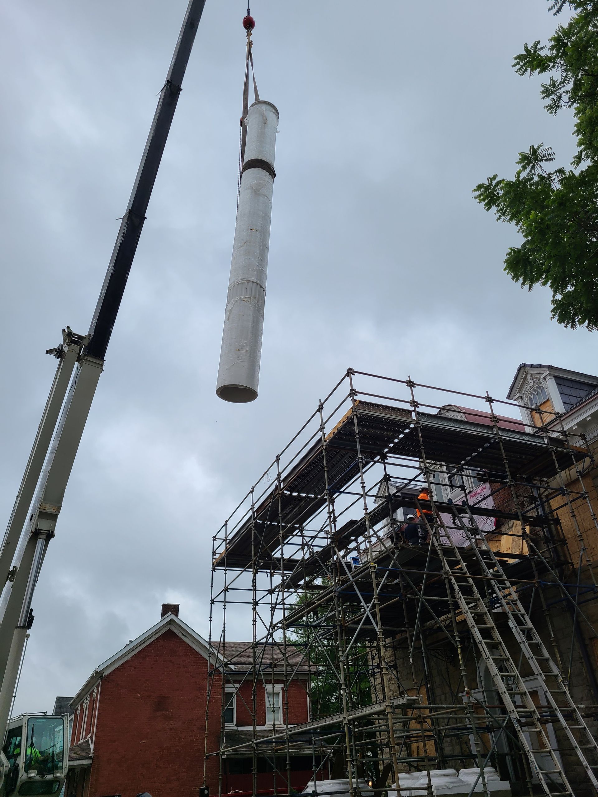 Crane lifting a long, white cylindrical object near a building with scaffolding on an overcast day.