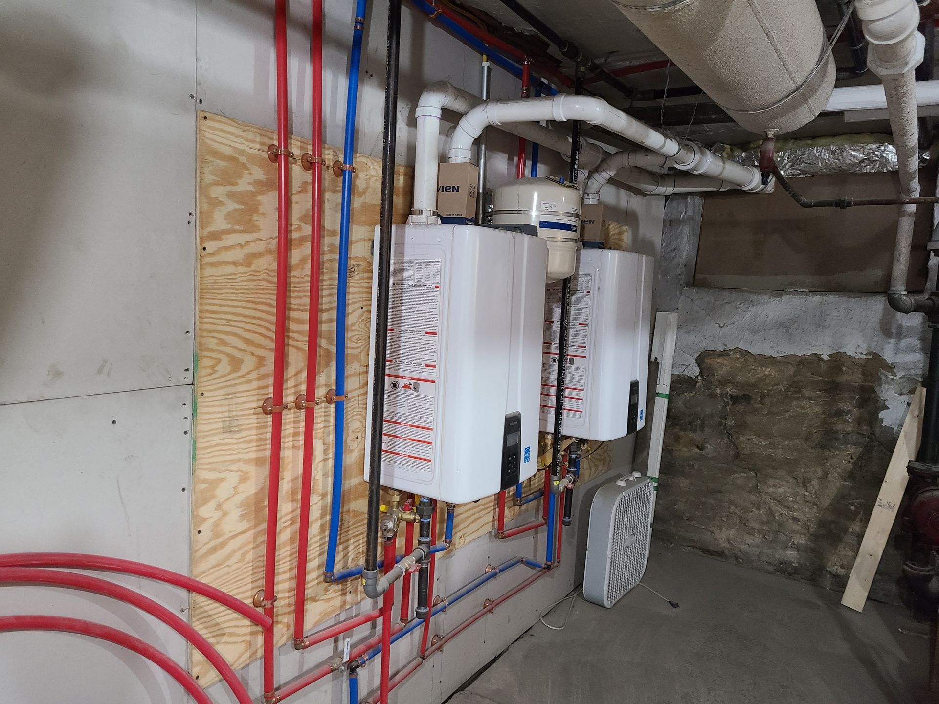Two white tankless water heaters mounted on plywood in a basement, with red and blue pipes.