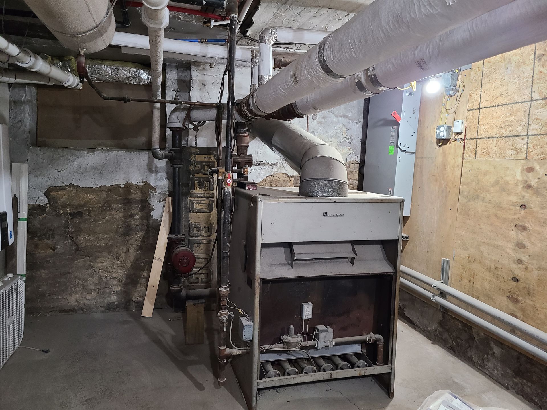 Furnace in a basement. Pipes overhead. Dark, aged brick and drywall walls.