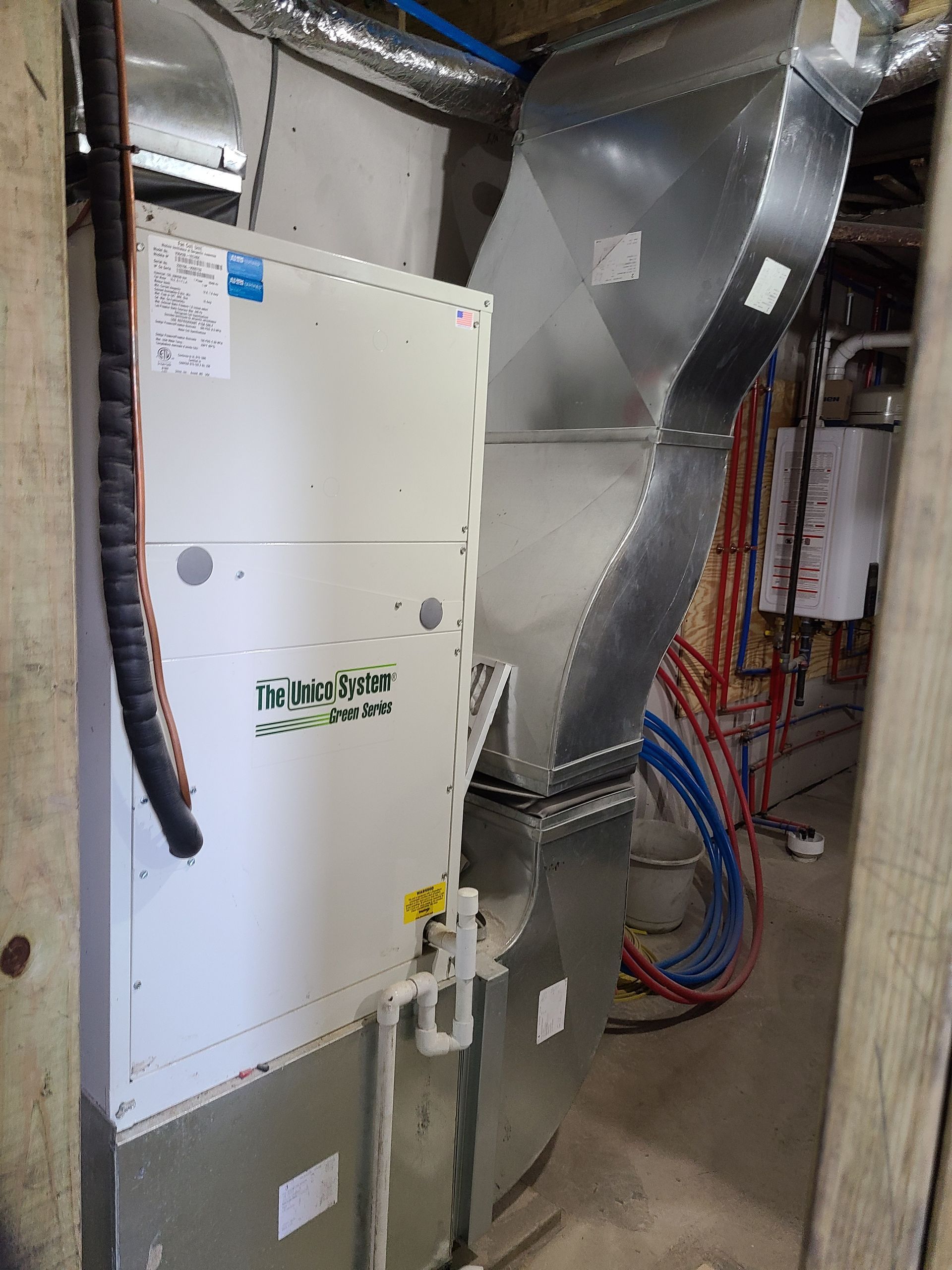 HVAC system with metal ductwork in a basement setting. White unit with green accents. Blue and red pipes visible.