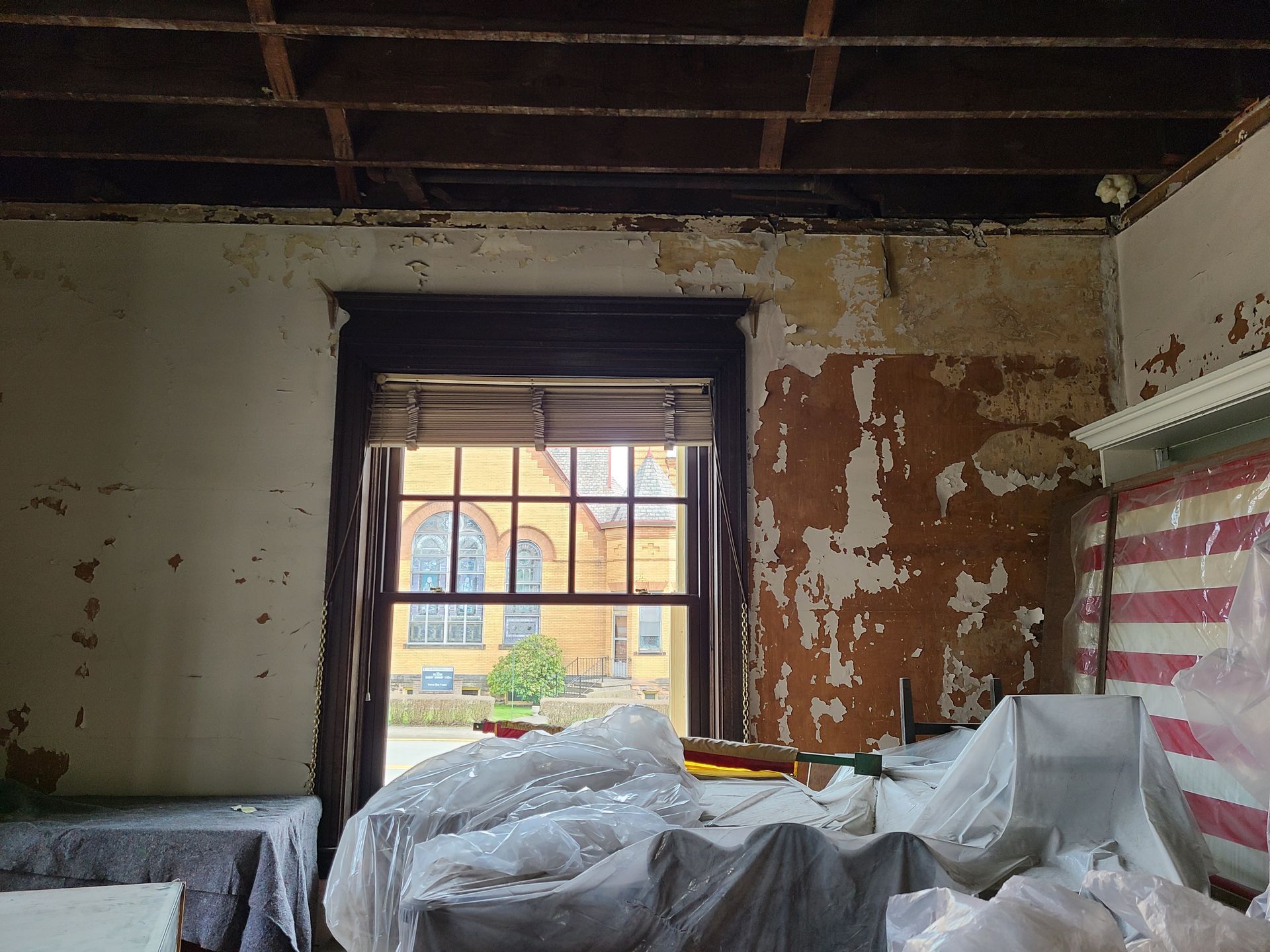 Interior view of a room with peeling paint, open window, exposed ceiling, and debris.