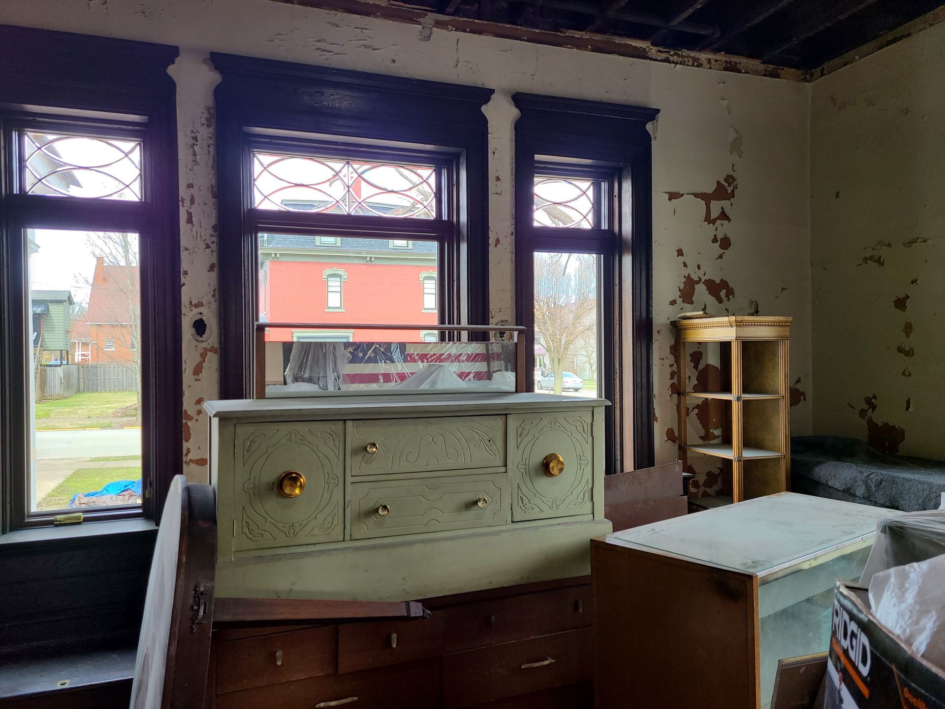 Room with peeling paint, three windows, antique furniture: dresser, shelving unit, and storage box.