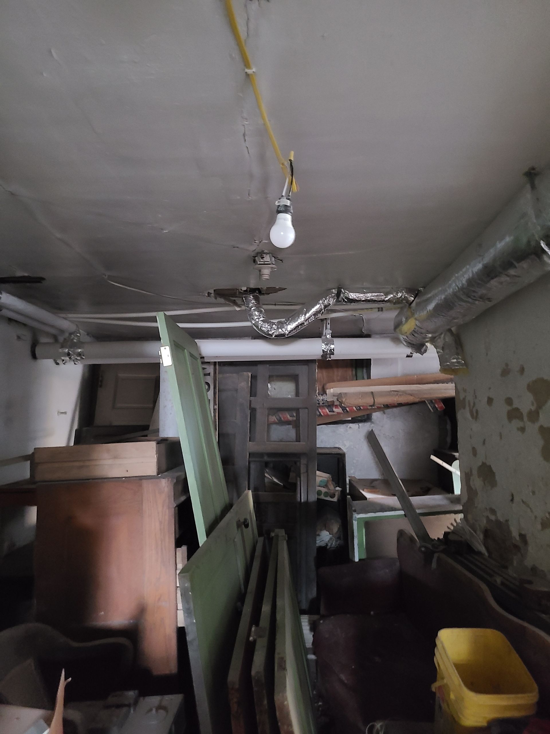 Cluttered, dimly lit basement with exposed wiring, exposed pipes, and debris.