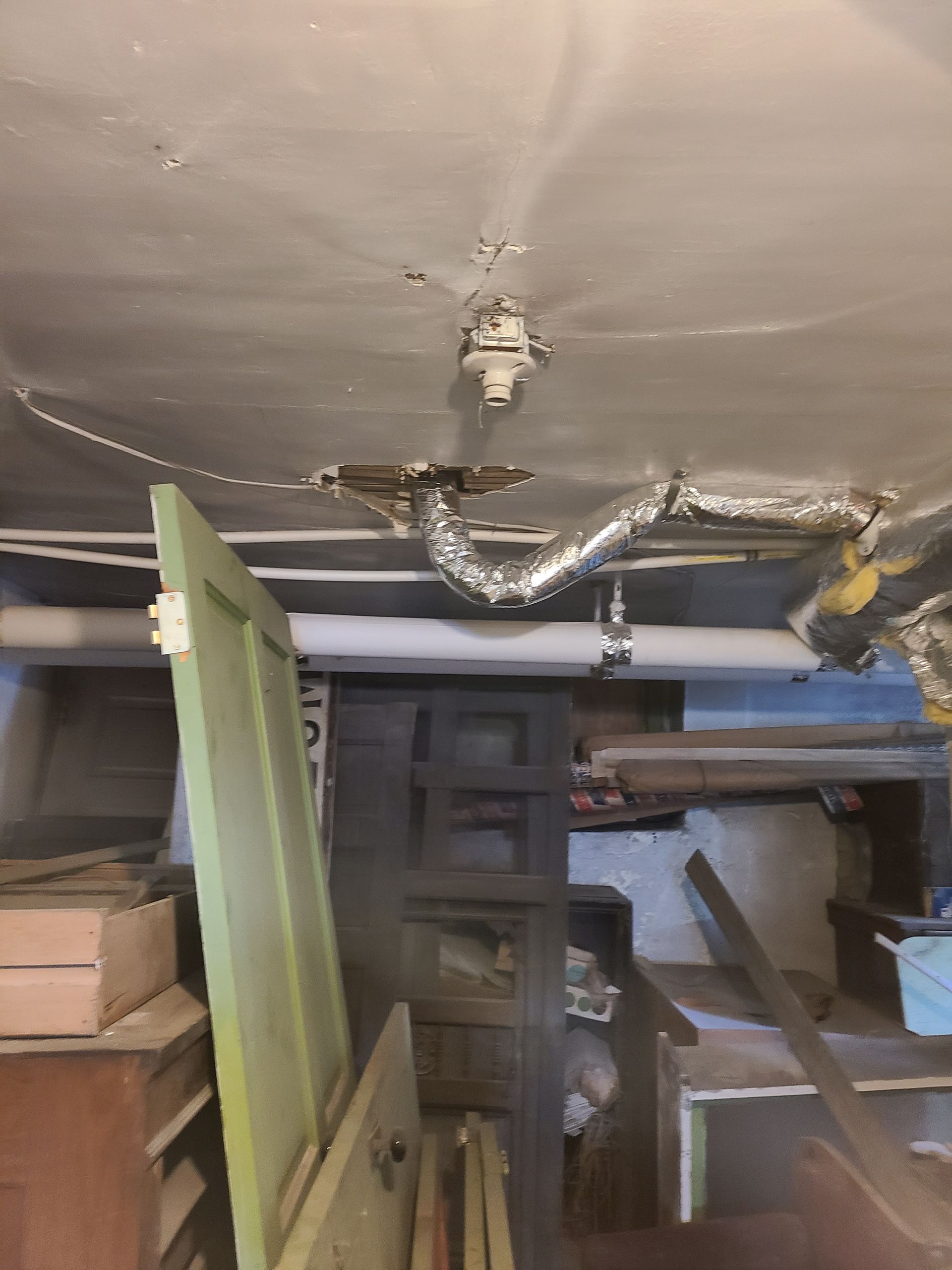 Basement ceiling with exposed pipes, ductwork, and light fixture. Green door and cluttered storage visible.