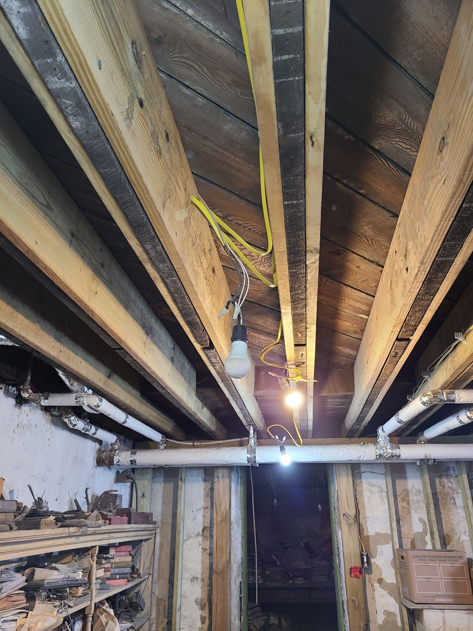 Wooden ceiling with exposed beams, pipes, and dangling lightbulbs in a dim interior.