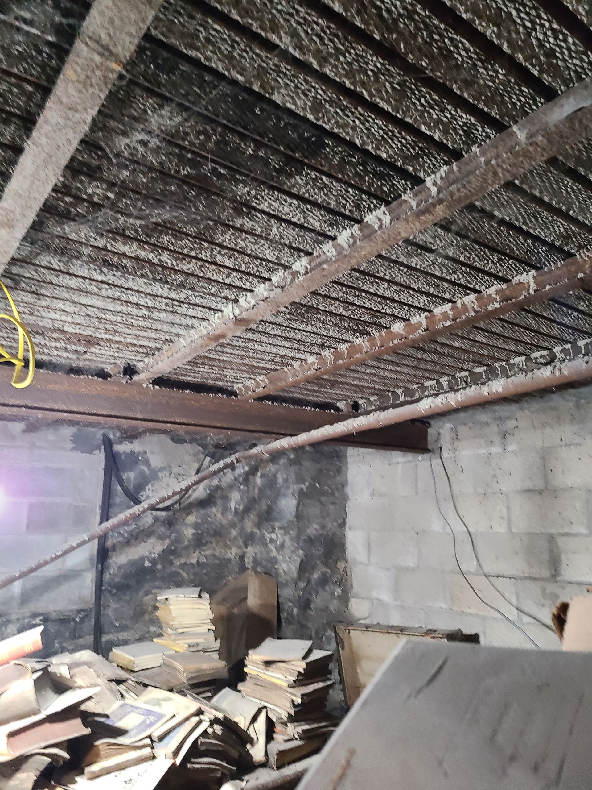 Basement ceiling with weathered wooden beams and accumulated debris. Concrete block wall and pile of materials below.