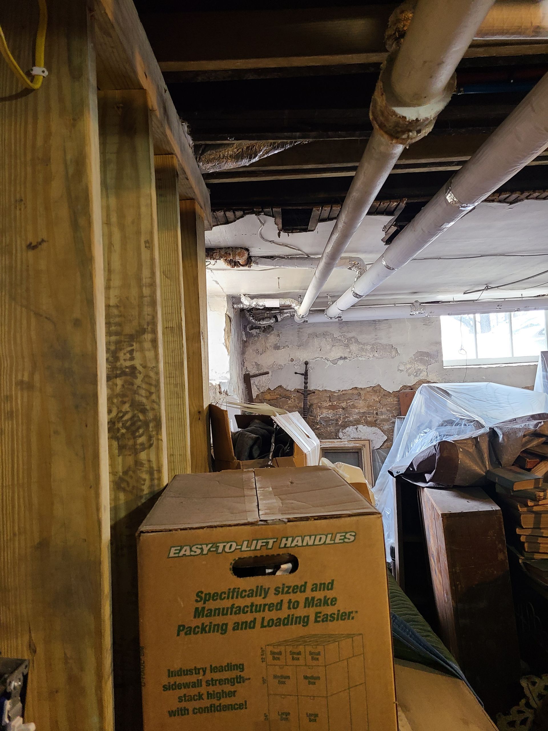 Basement interior with exposed beams, pipes, and cardboard box. Brick wall and small window visible in the background.