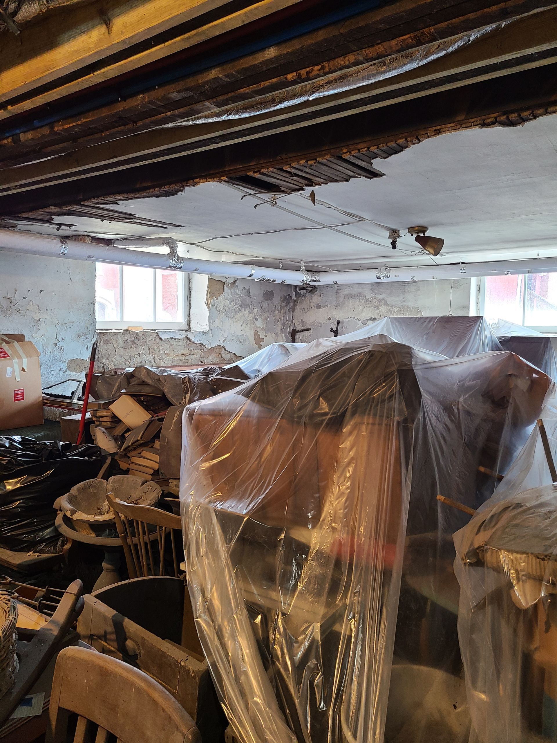 A cluttered basement with exposed ceiling beams and plastic-covered furniture. Windows and debris are visible.